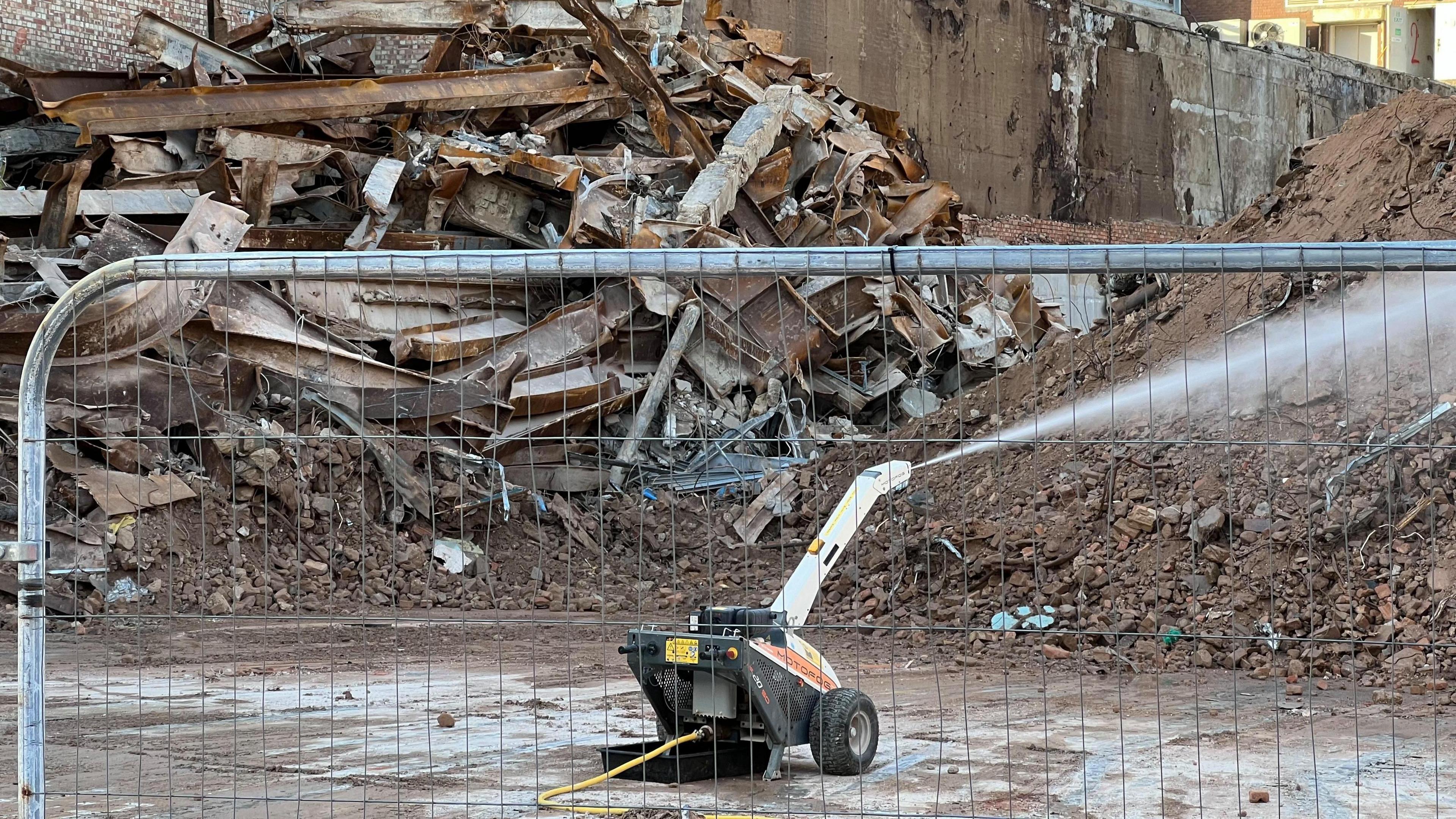 A small black and white robotic machine spraying water on rubble, with a high pile of rubble in the background.