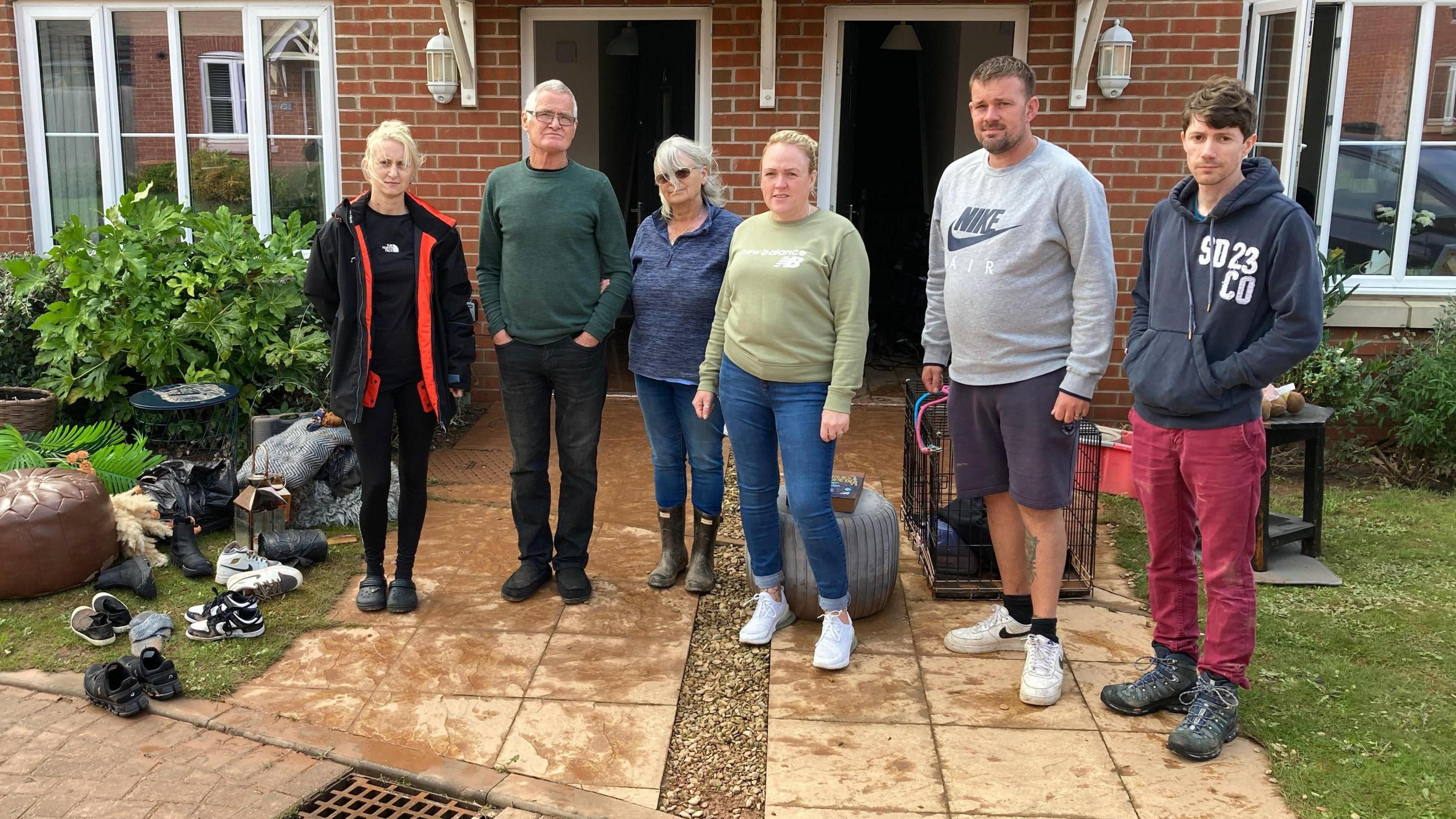 Three men and three women stand outside a pair of semi-detached houses with damaged household goods on the lawn and drive around them.