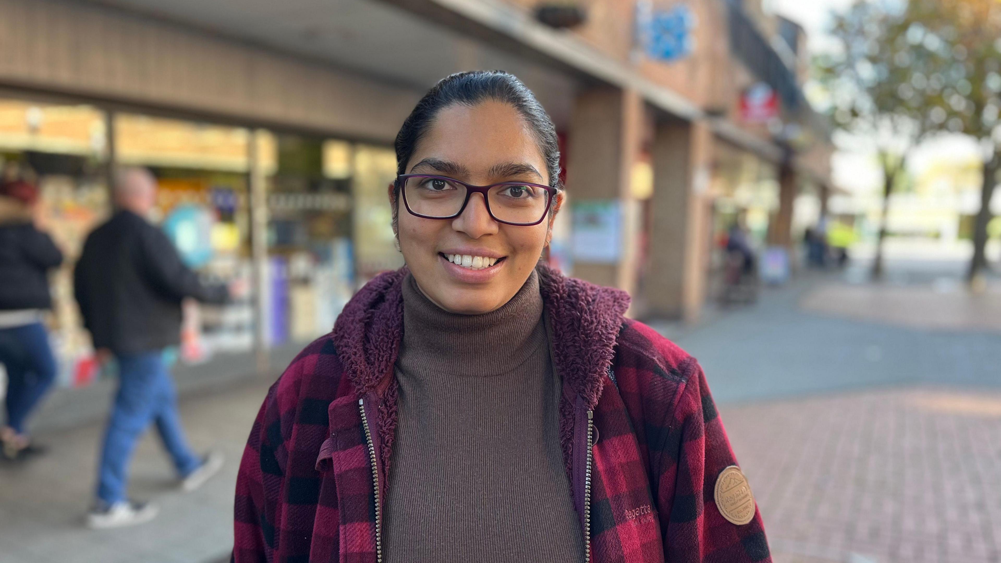 Aparna Valsala wears glasses, a red fleeced jacket and a rollneck jumper. She is smiling while standing in front of the Co-op store