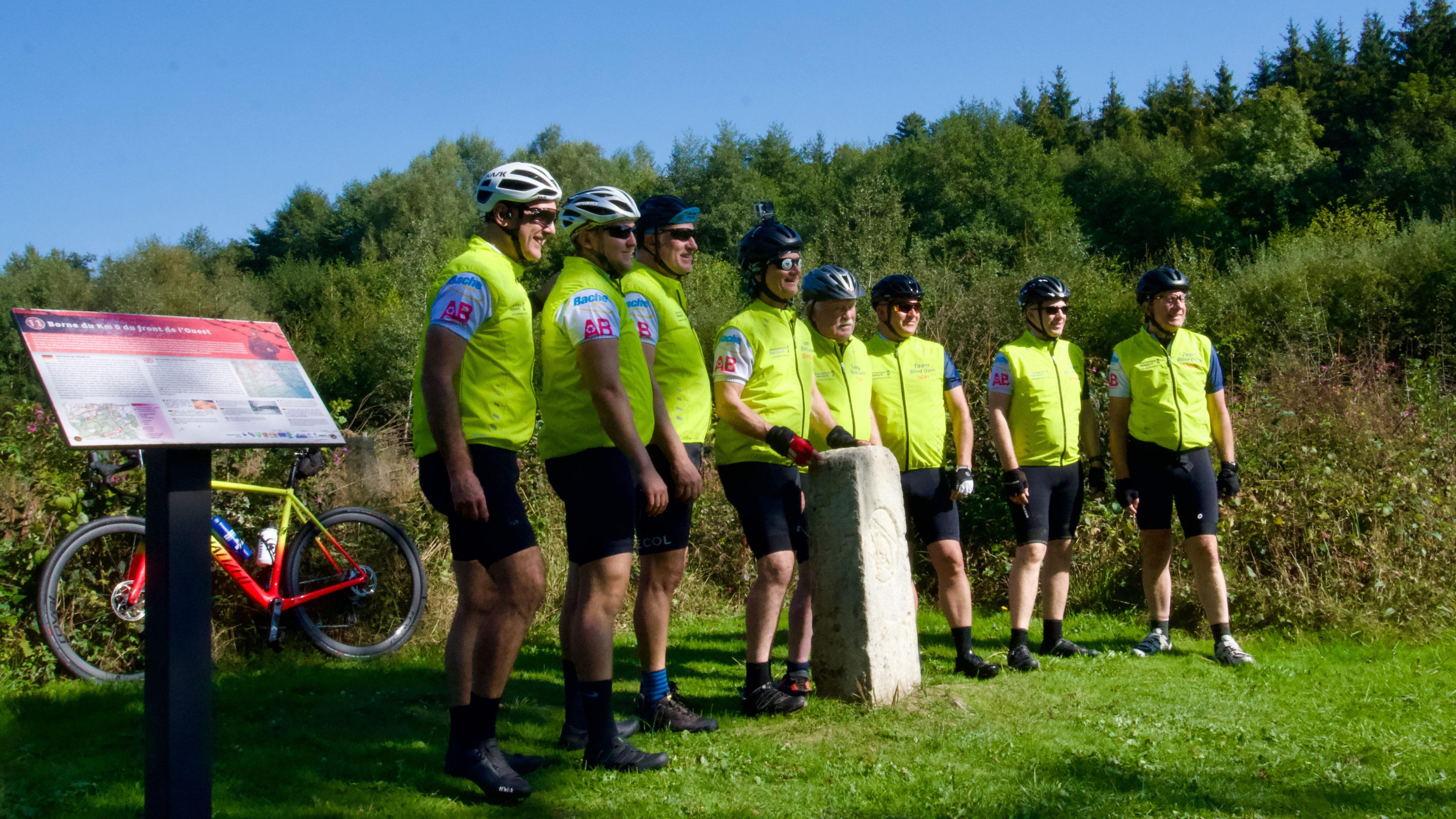 Eight cyclists in lime green gilets and cycling gear, standing in the sunshine at kilometre zero in Switzerland 