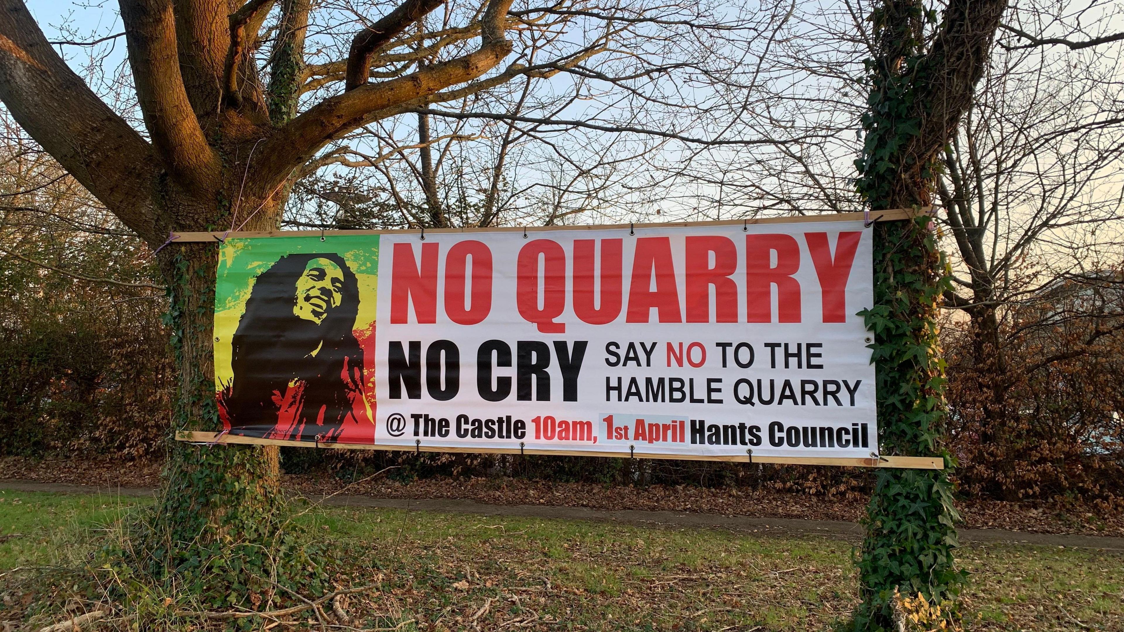 A large banner has been hung up between two trees in the countryside. There is hedging in the background and the tree trunks are covering in trailing ivy surrounded by grass. The banner has a silouhette black image of Bob Marley printed on top of the Jamaican flag colours of green, yellow and red. To the right of his image, the banner reads 'No quarry, no cry. Say no to the Hamble Quarry @ The Castle 10am, 1st April Hants Council'