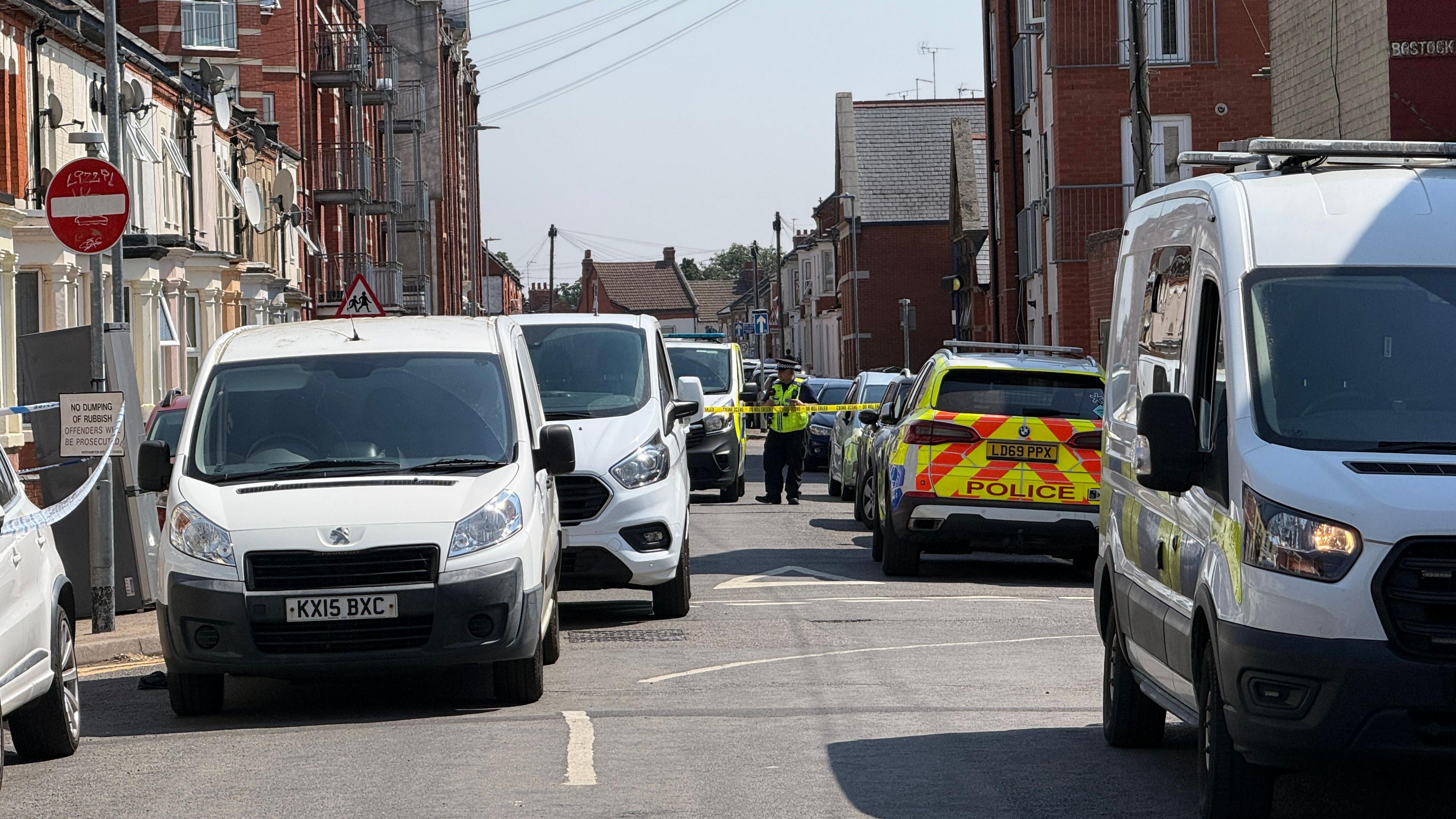 A late Victorian-style terraced street of houses with cars and buildings on either side. There are a lot of white vans on the road, a cordoned-off area, with a police officer standing in the road. Road signs are to the left. 