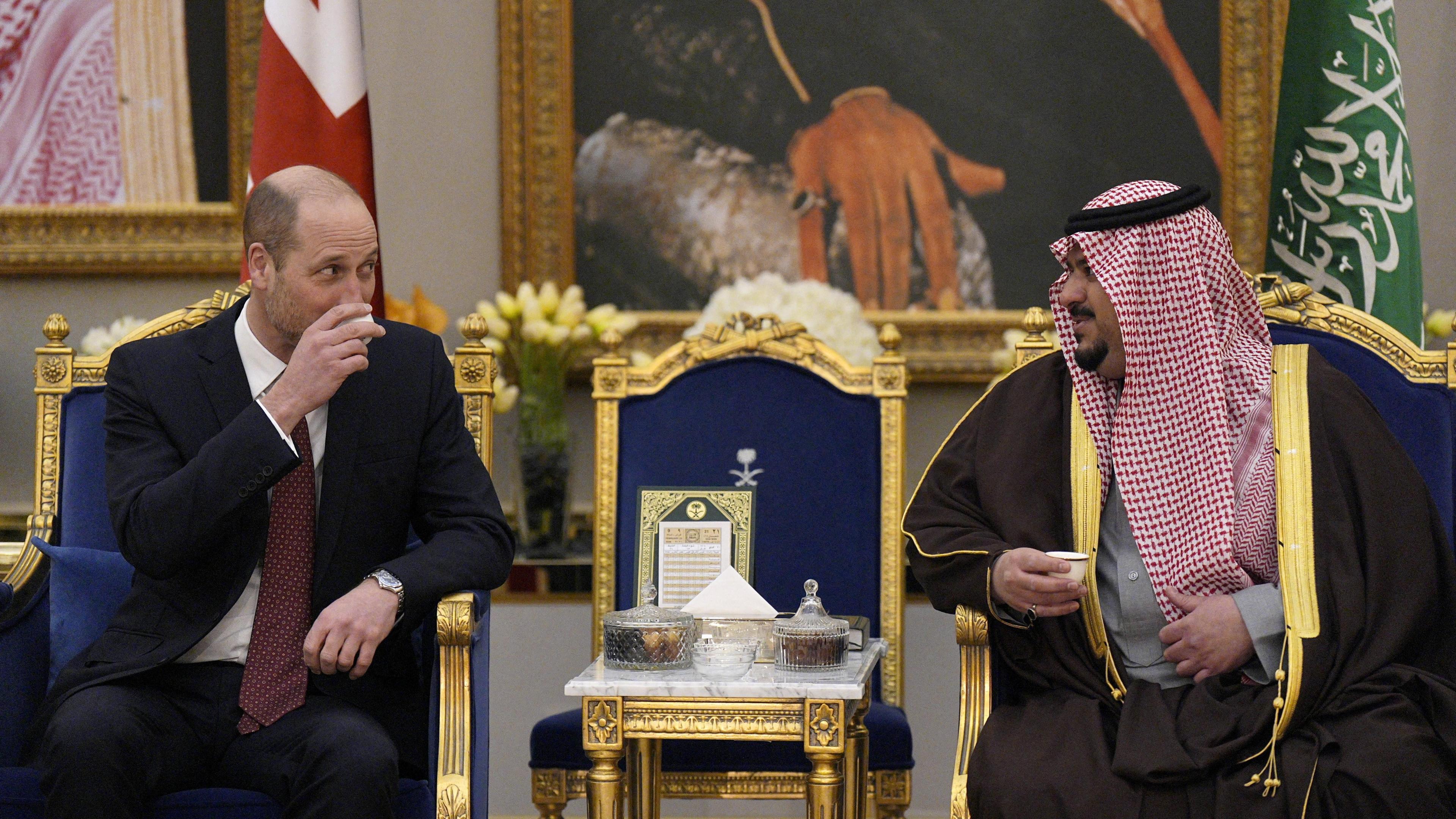 Princi William ulet në një karrige blu dhe të artë dhe pi nga një filxhan i vogël qeramik me Zëvendësguvernatorin e Rajonit të Riadit, Princin Mohammed bin Abdulrahman bin Abdulaziz.