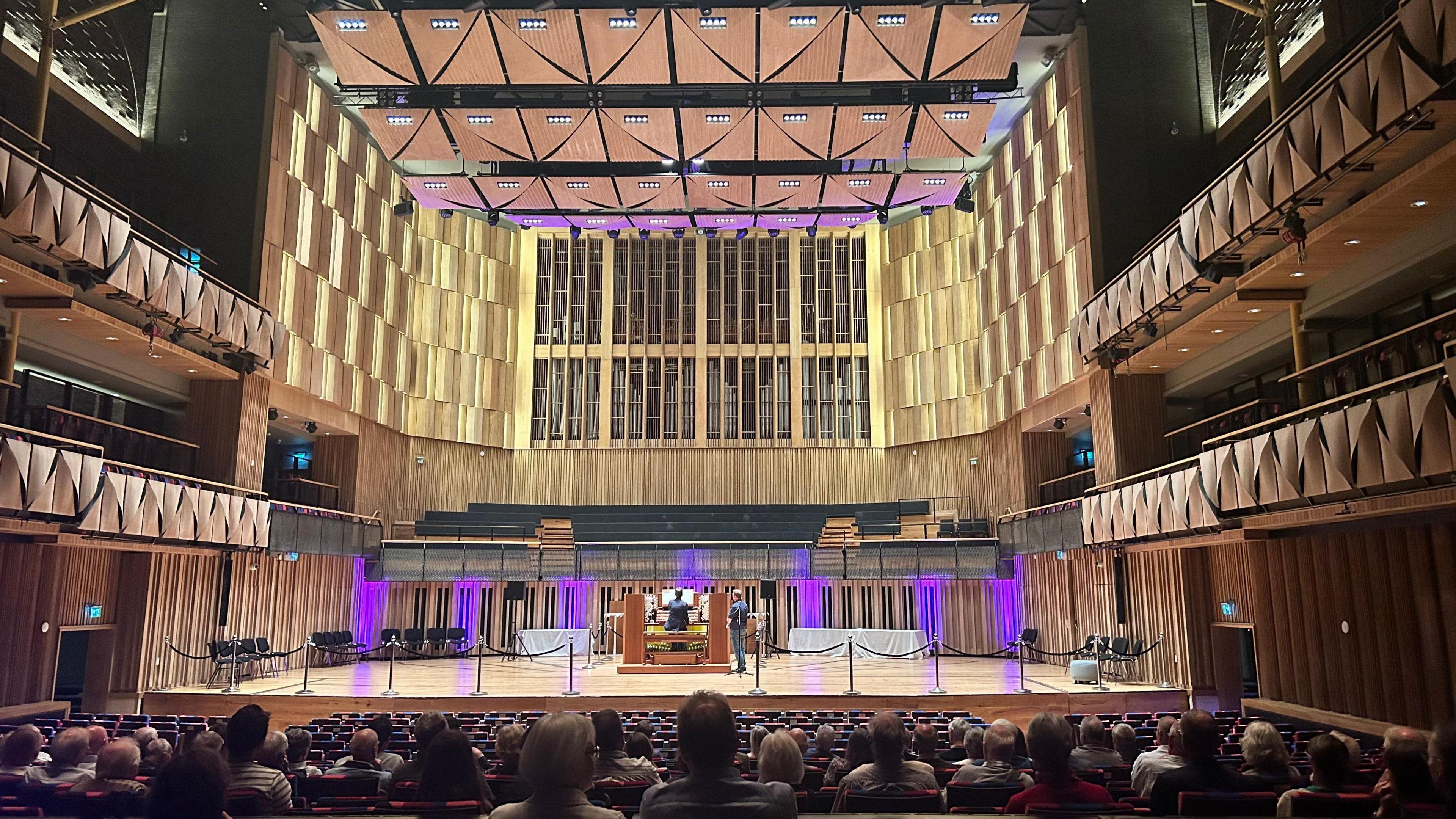 Image of a concert venue, with a seated audience in the foreground watching a man on stage playing a vast pipe organ