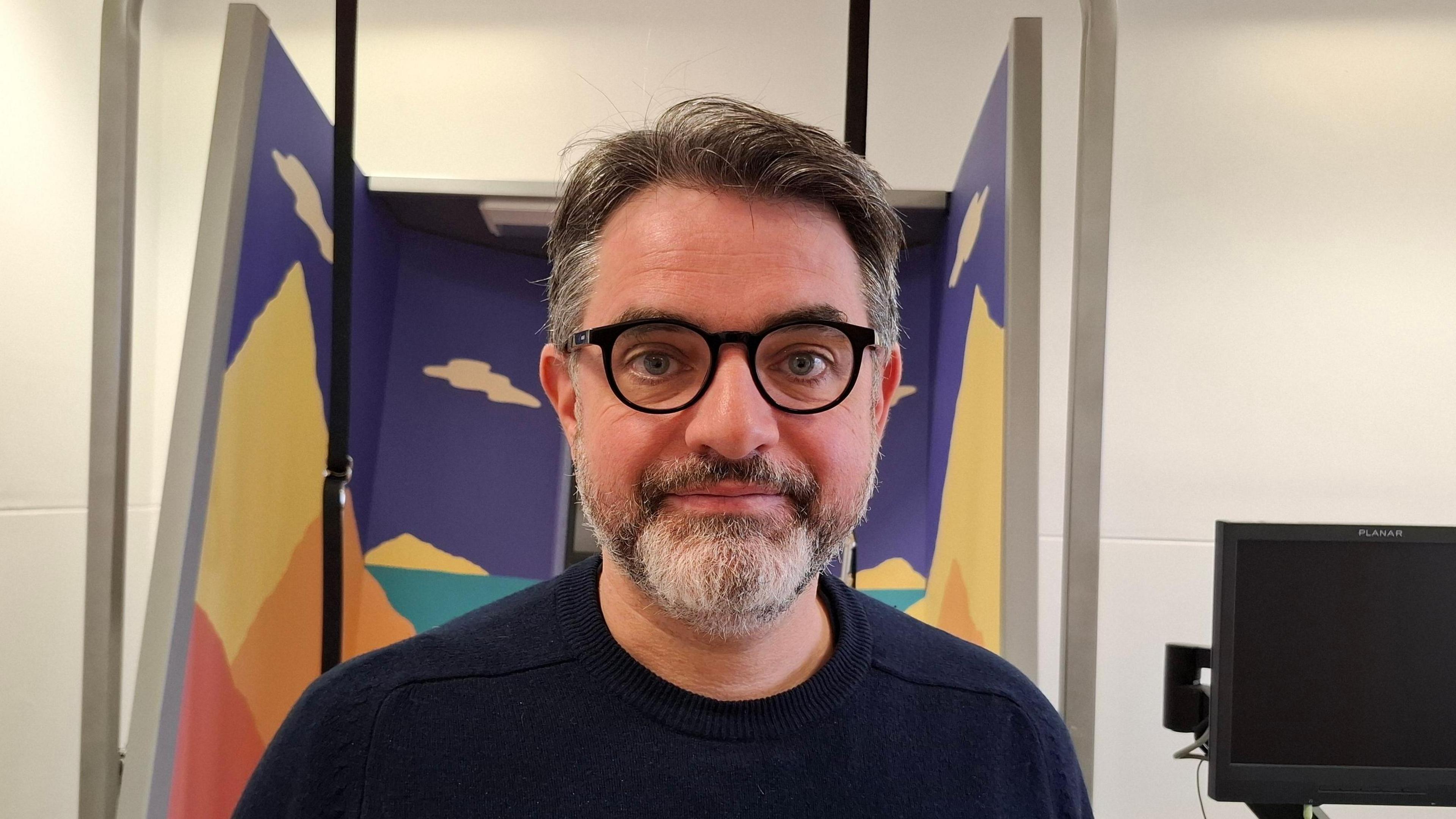 Close up image of a man with black glasses and black jumper who is smiling at the camera