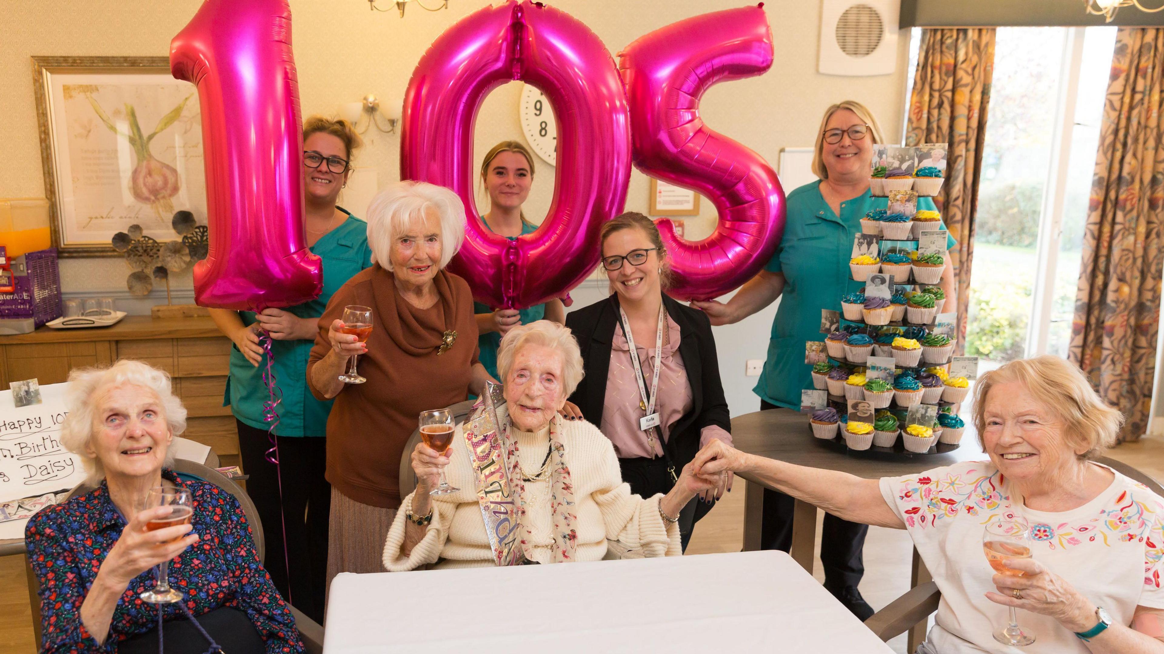 Ms Taylor sat at a white table surrounded by seven women. Balloons making the number 105 are in the background. A tier of cupcakes is to the right hand side.