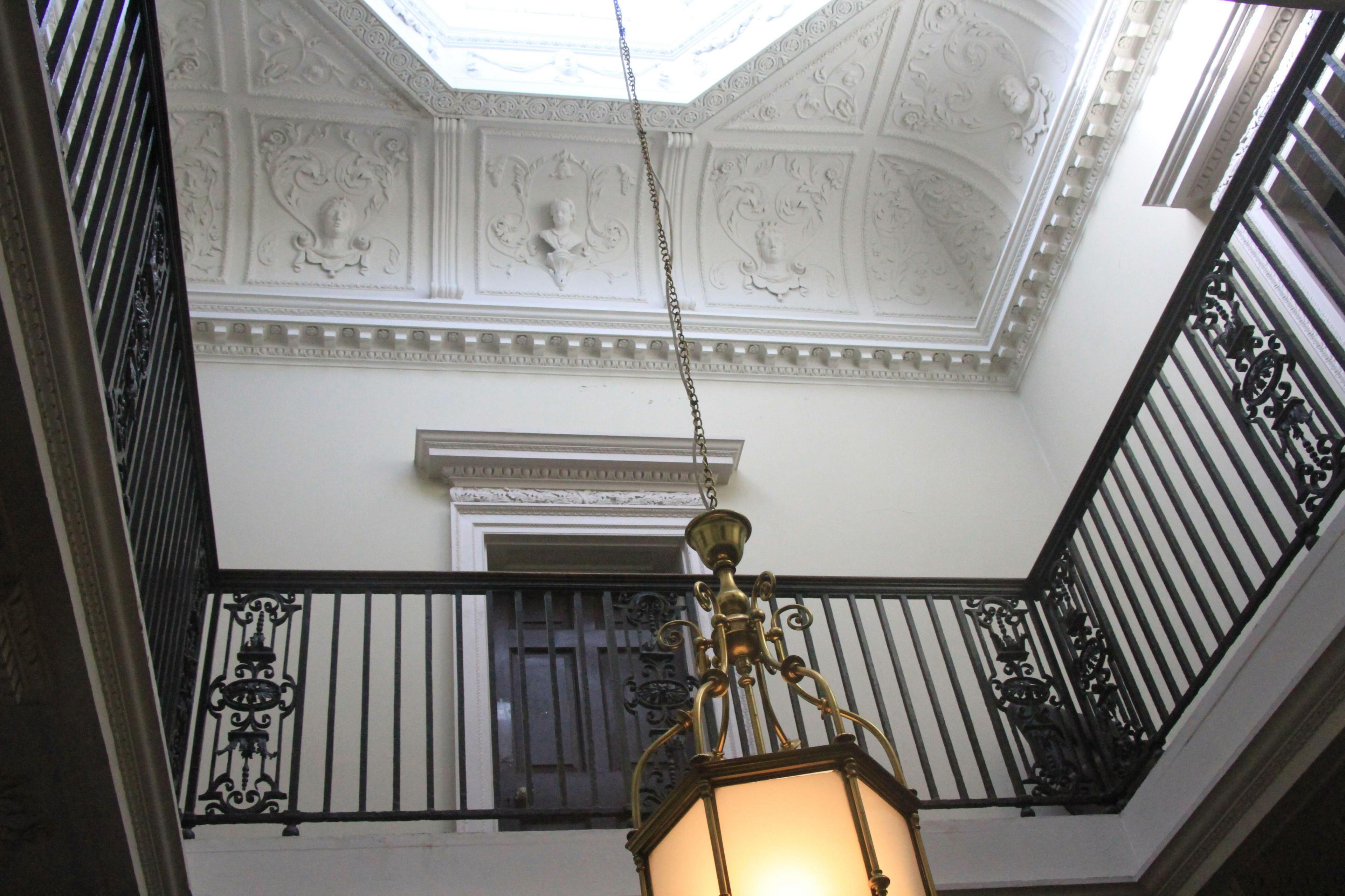 An ornate entrance hall consisting of white walls and ceiling. A mezzanine floor is visible with black iron railings. A large lantern hangs in the centre of the hall.