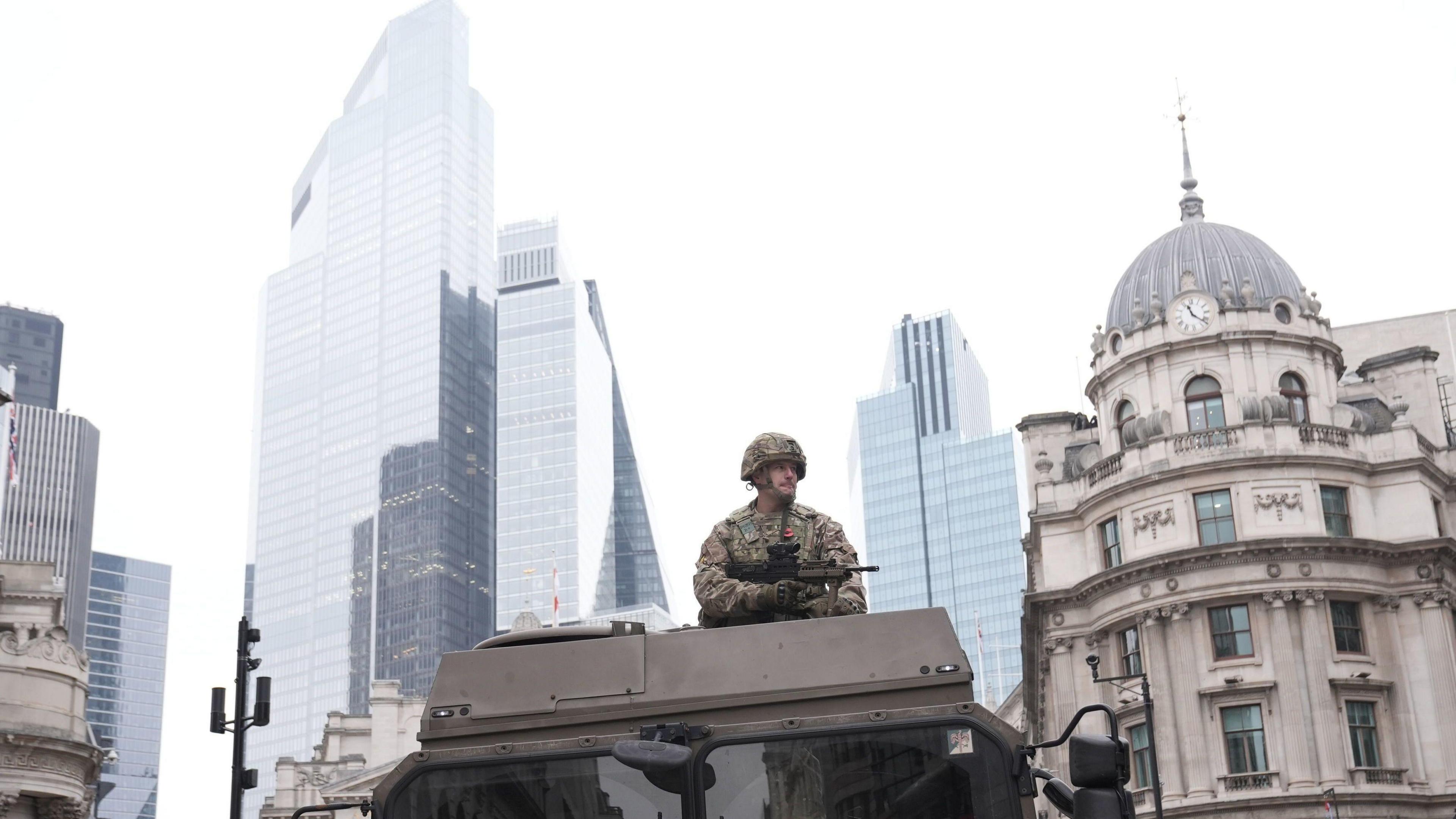 A soldier stands atop an armoured military vehicle holding a weapon, set against the backdrop of modern skyscrapers and historic architecture in the City of London.