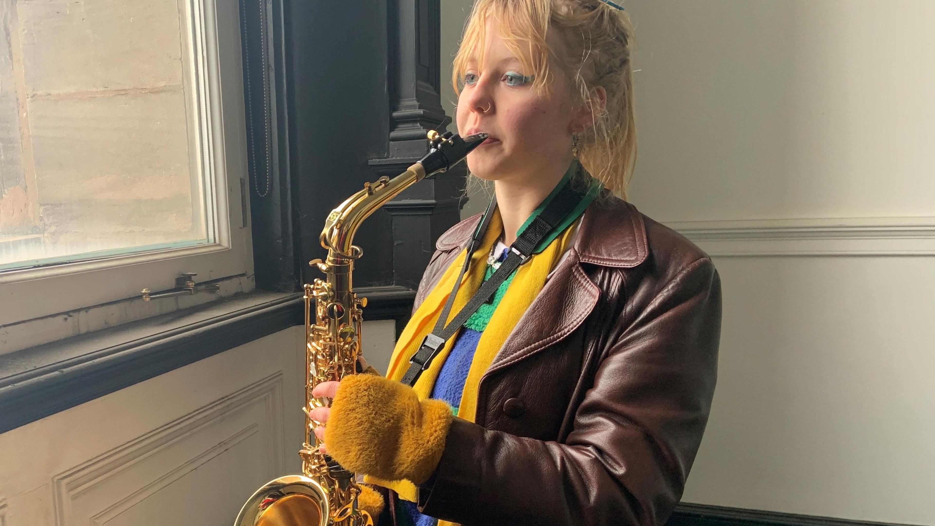 Adha Charles has blond hair tied back in a pony tail, she is wearing a brown leather jacket with a yellow scarf and yellow gloves. She is stood by a large upstairs window playing the saxophone