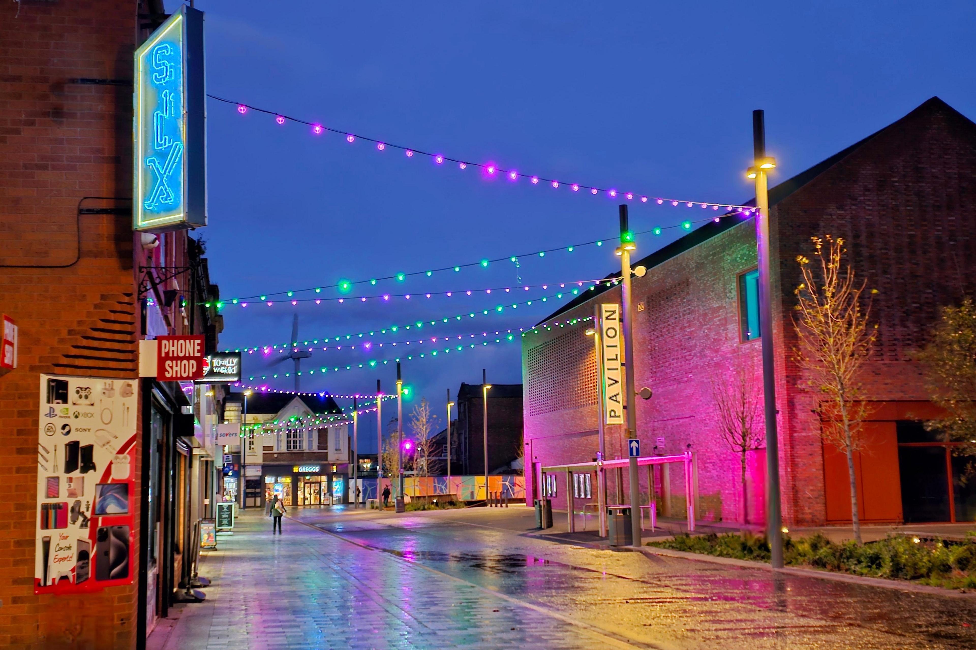 Green and pink Christmas lights are attached to lampposts and criss-cross over a street. Lit shop signs also illuminate the street.