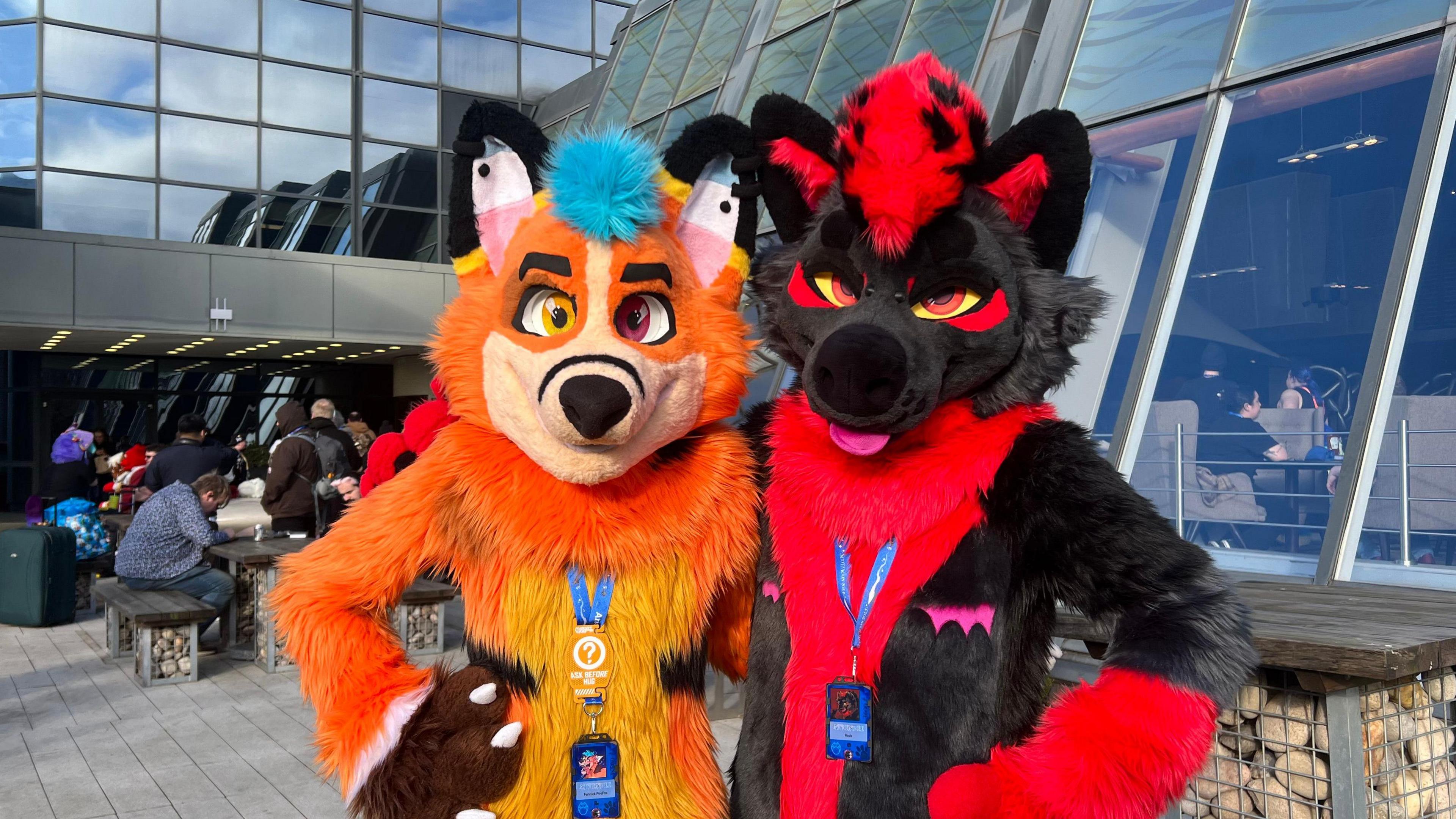 A person in a large orange fox costume stands next to someone in a large red and black dog costume. 