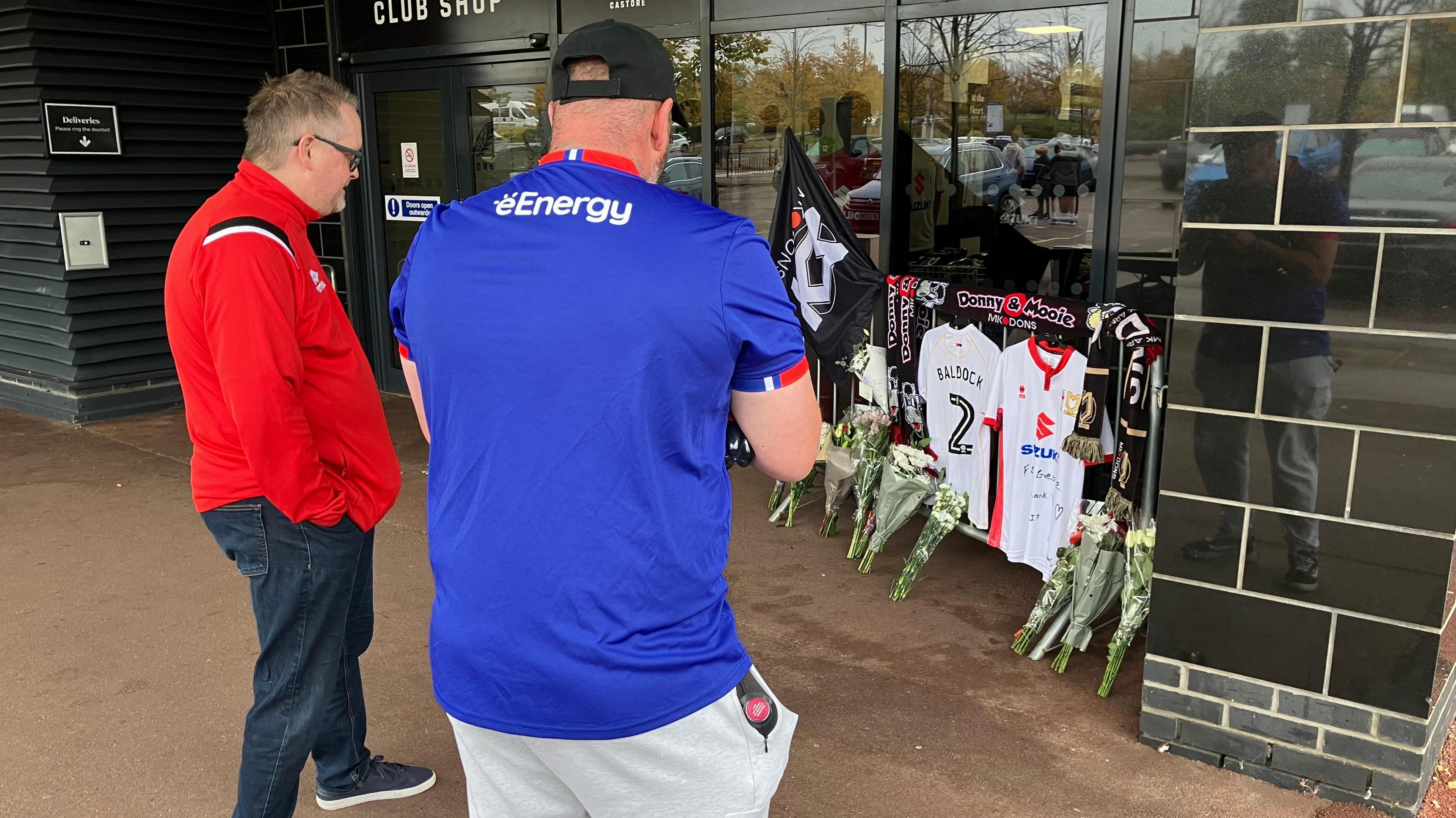 Fans gather at exterior memorial outside club entrance to late George Baldock. Football shirts and flowers can be seen. Two men take photographs.