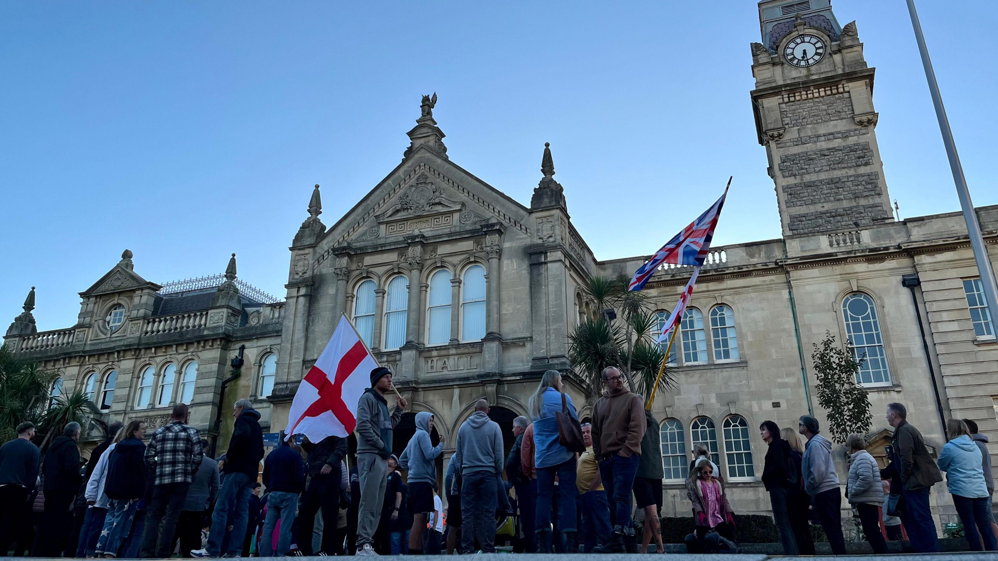 A group of protesters, one holding a St George's Cross and another holding a Union Jack, stand outside Weston Town Council. It is growing dark, though the sky is still blue and the impressive stone building is still clearly visible. 