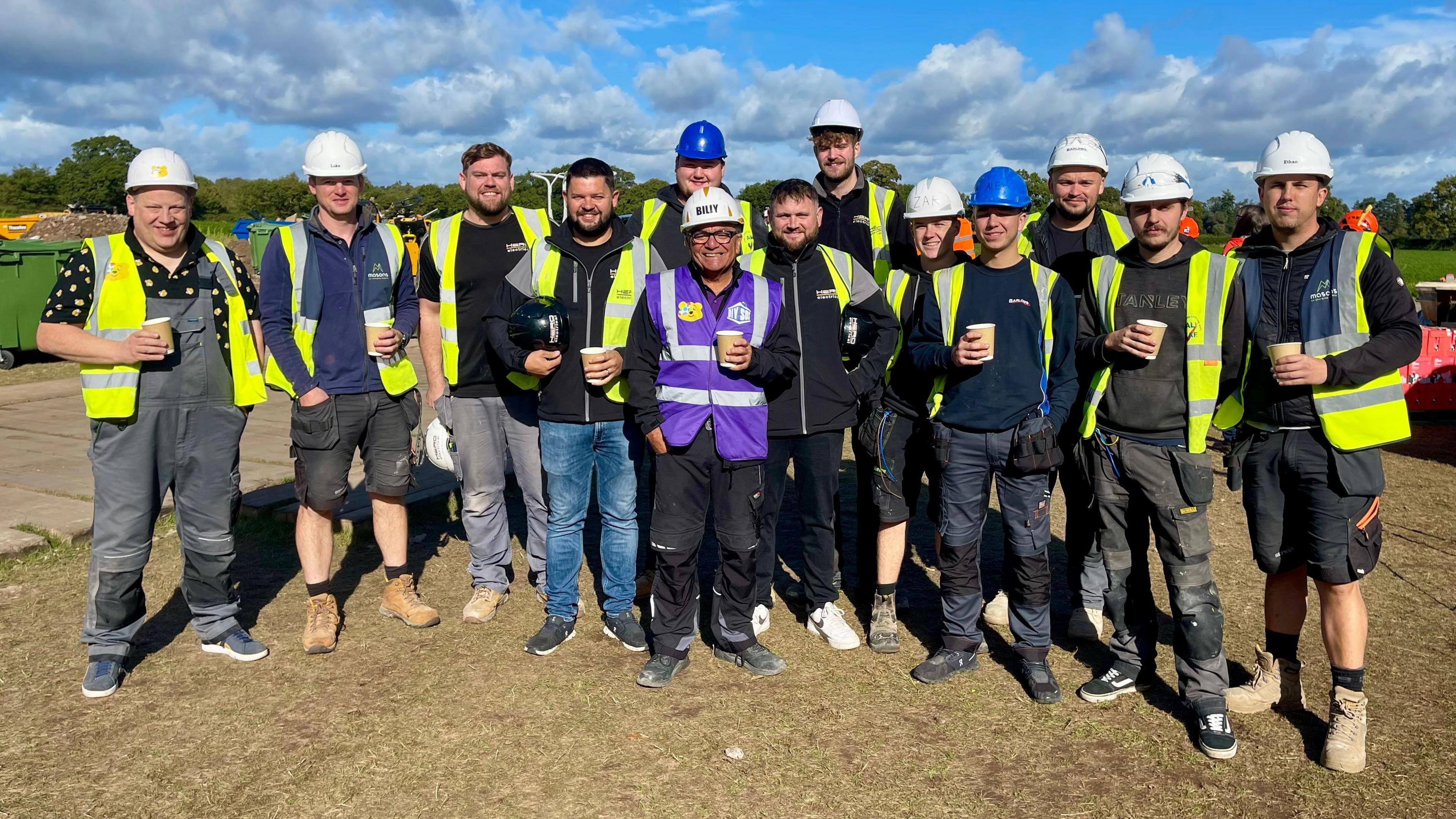 Thirteen men dressed in hard hats, yellow hi vis vests and workboots stand together on a worn grassy area. They all wear a variety of workwear suitable a building site. The man in the centre is wearing glasses, a purple hi vis vest and 'BILLY' is printed on his hard hat. The majority of them are holding disposable cups.