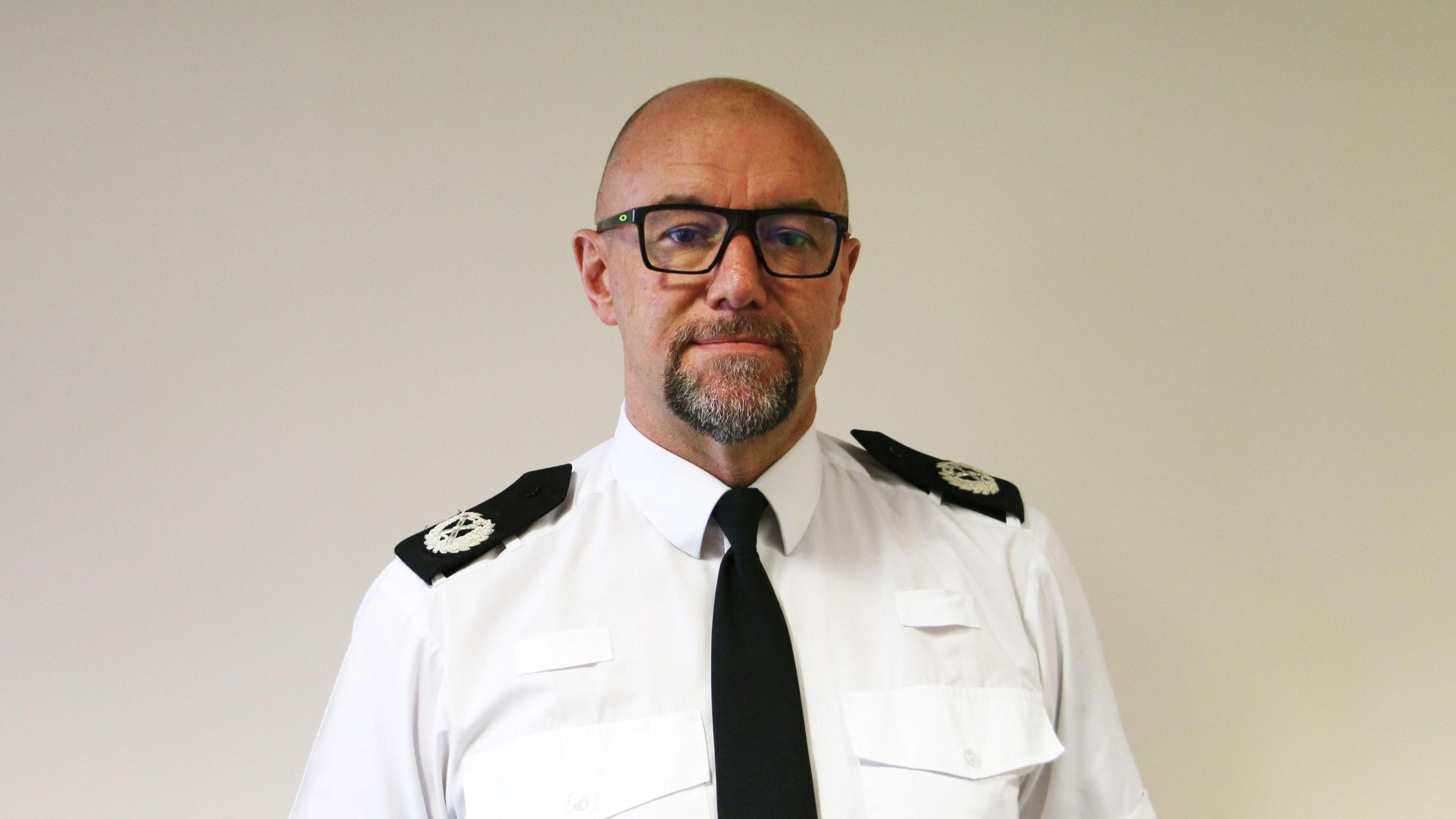 Man with square black glasses, grey beard and smooth head in a white police shirt with crests on his shoulders. His expression is neutral as he looks into the camera.