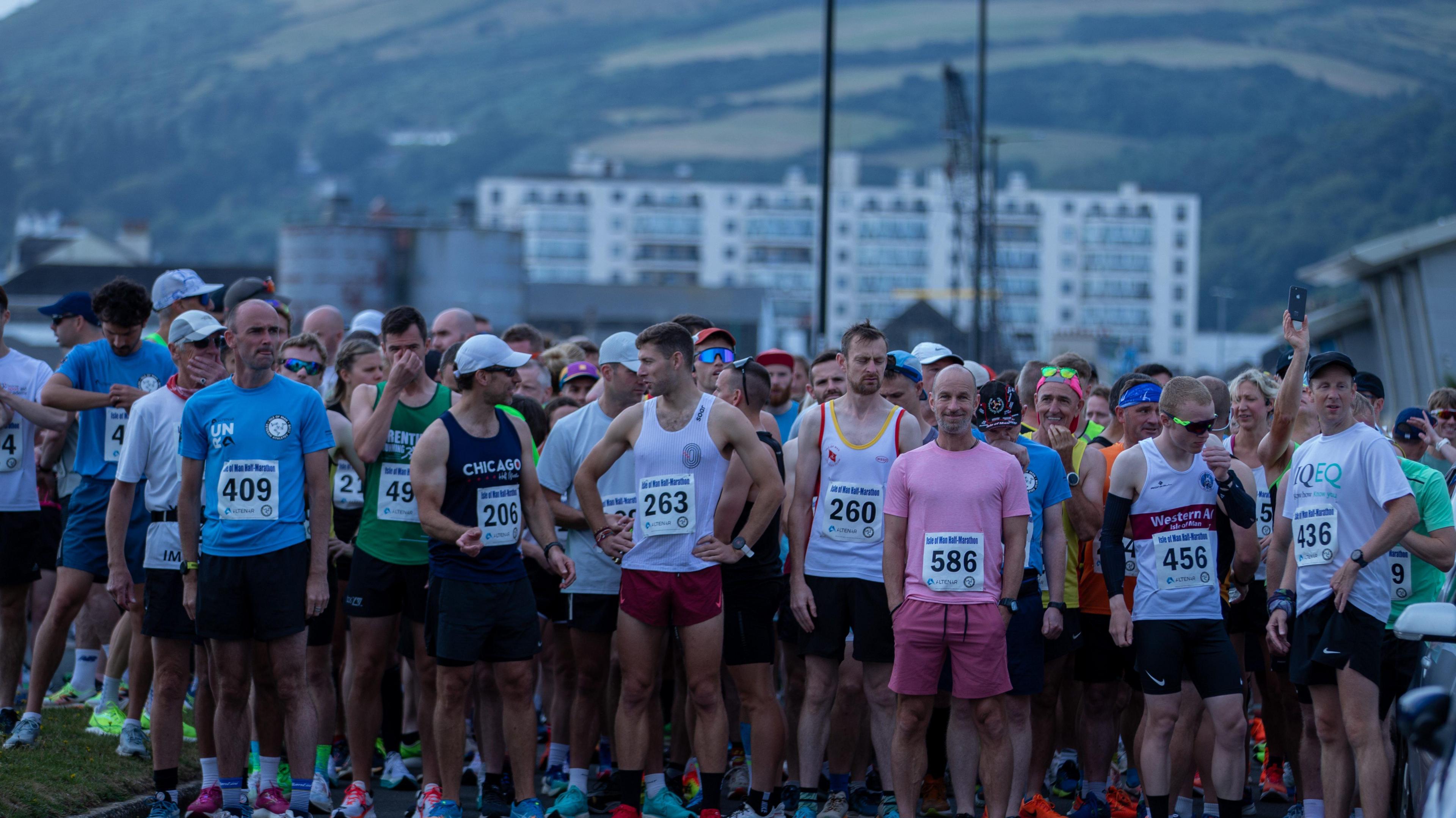 A large crowd of runners stand at the start line each has a number pinned to them, and you can see a buildings and a hill in the background.