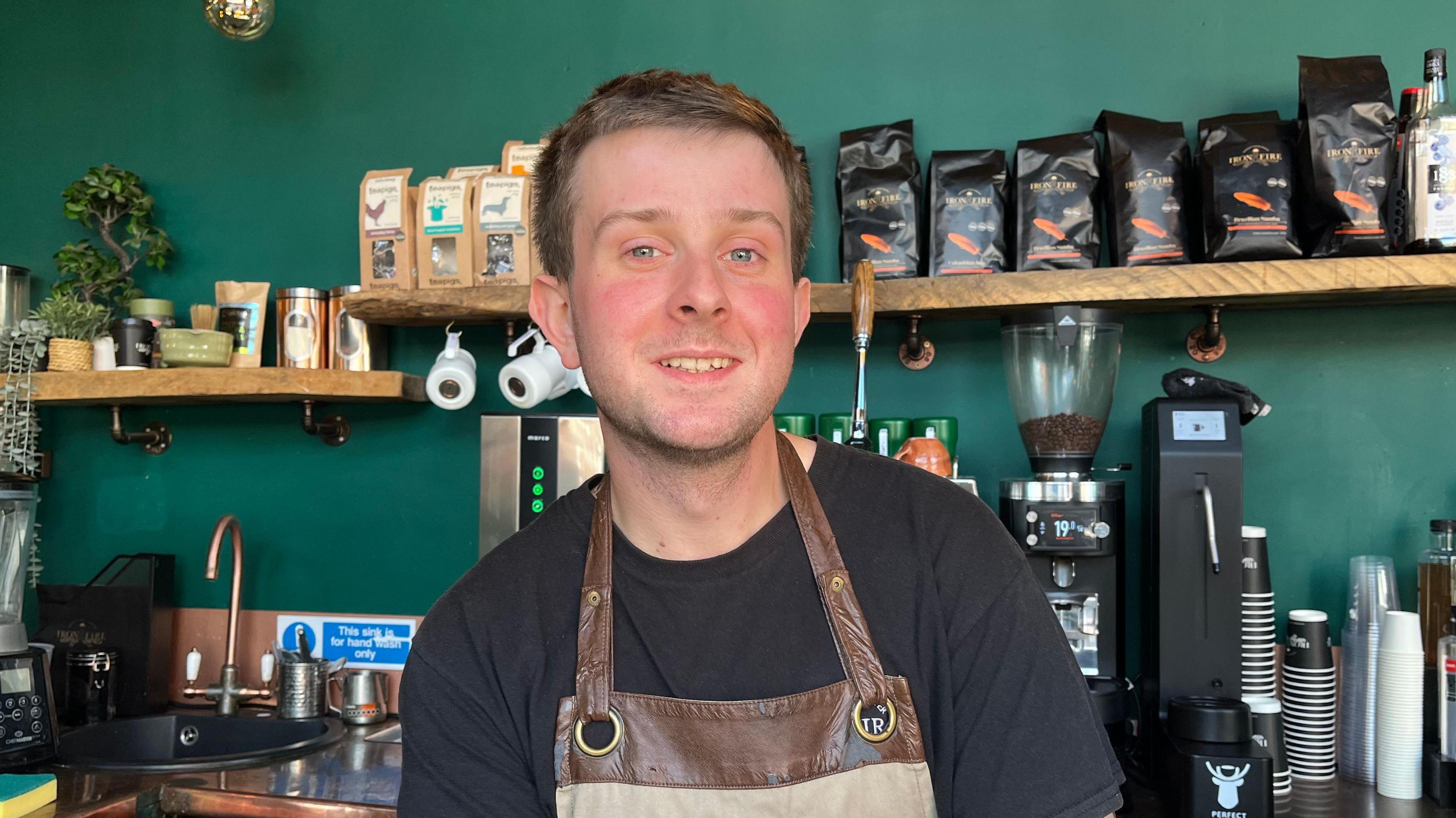 Nathan pictured smiling and looking into the camera. He's wearing a black tshirt and a brown leather apron. Behind him is a wall painted dark green, with wooden shelves with black bags of coffee on them. On the countertop below there are coffee machines, stacks of takeaway cups, and a brass sink.
