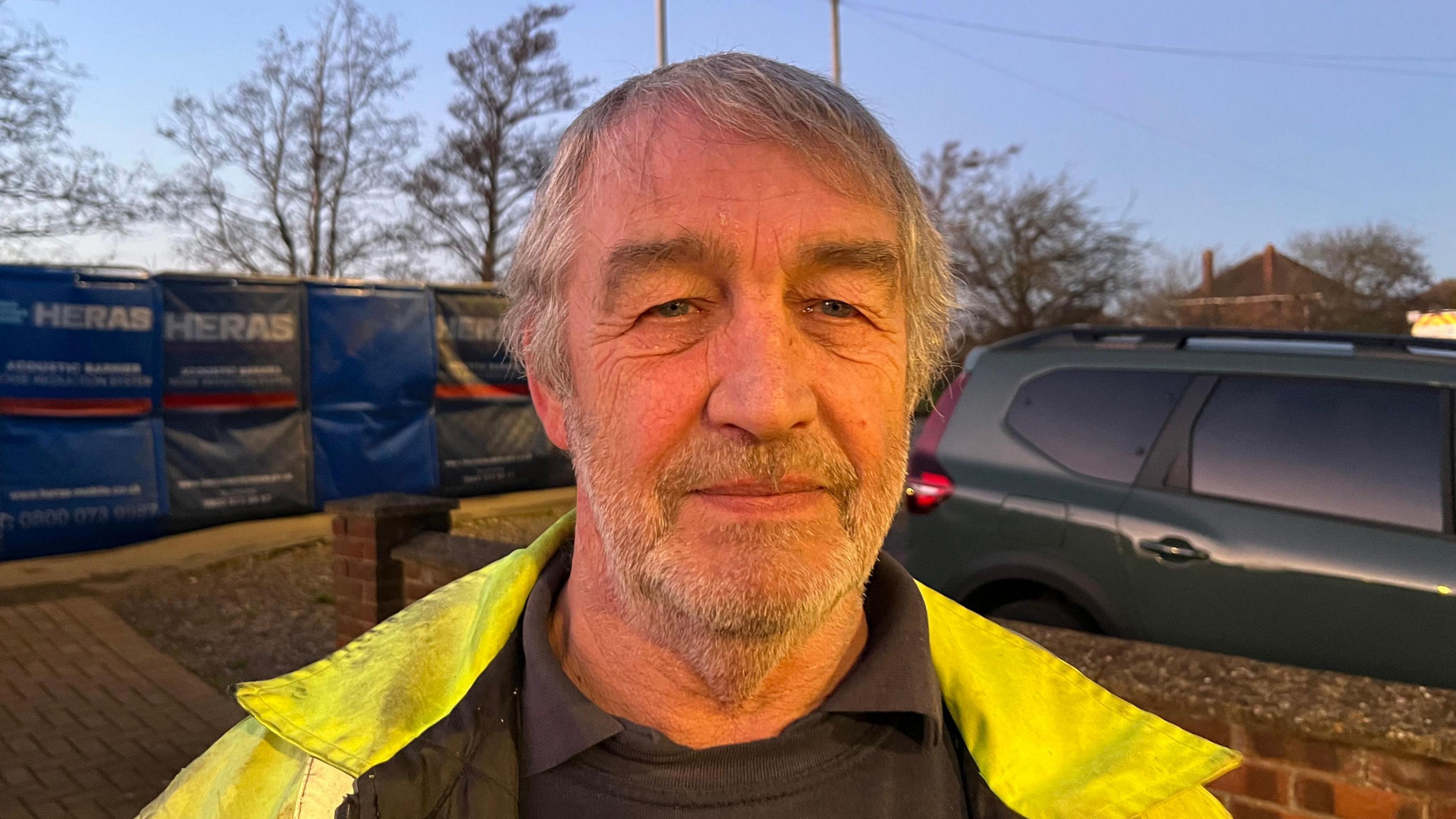 Mark Jackson looking into the camera on a driveway. He has short grey hair and stubble and is wearing a hi-vis jacket and black jumper.