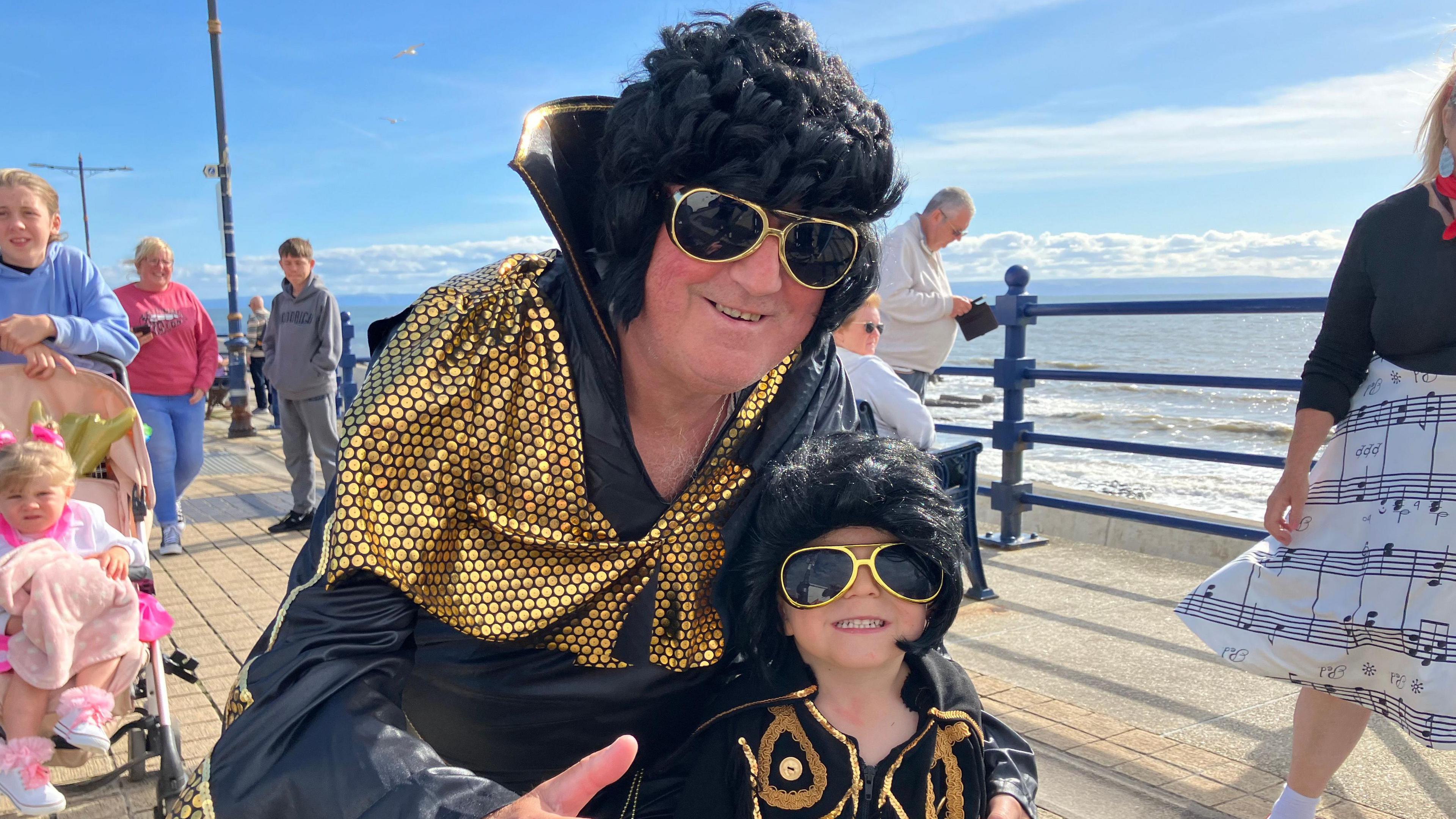 A man and a boy in matching Elvis outfits and spectacles smile 