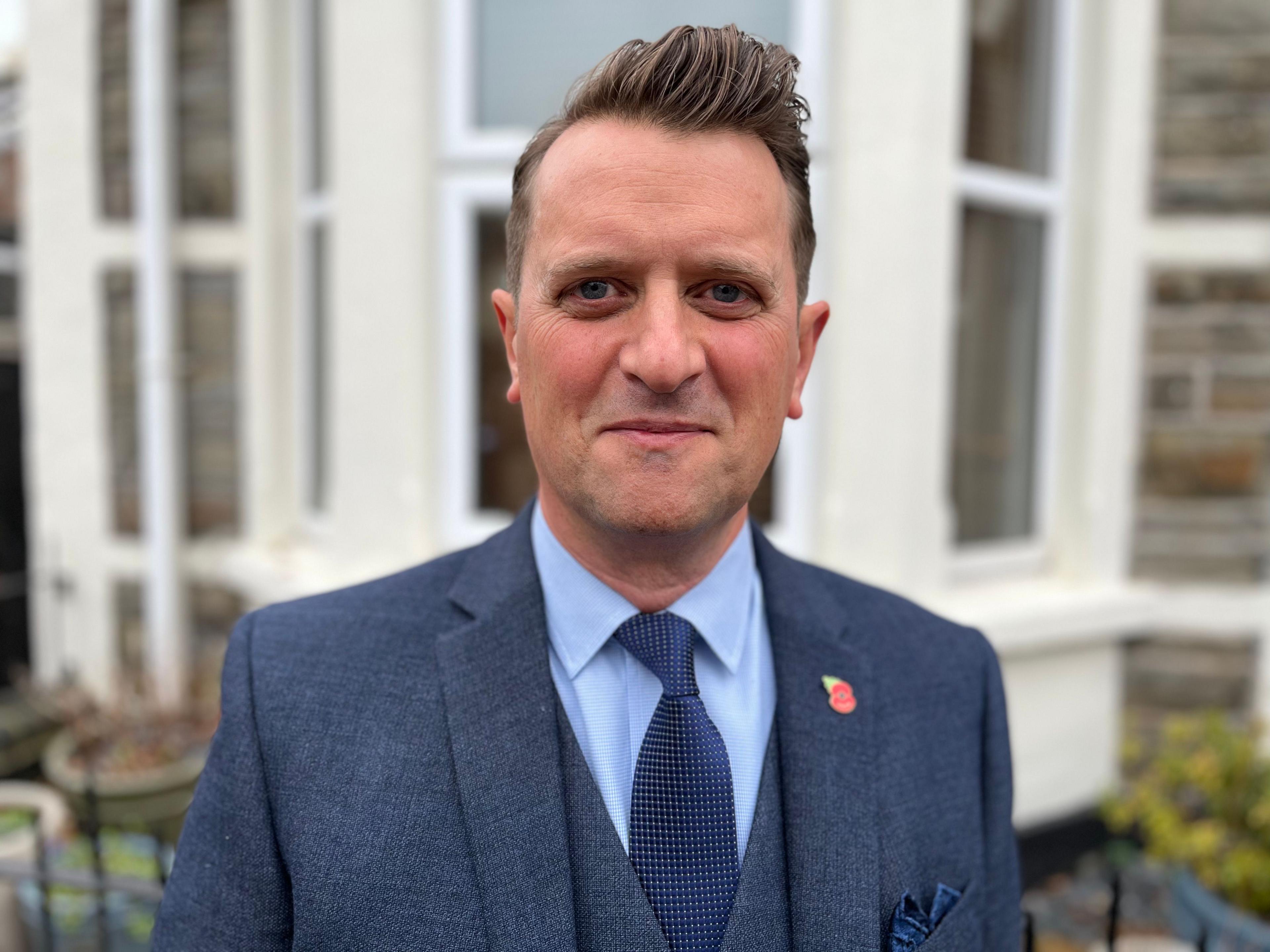 Historian Jeremy Banning. He is wearing a light blue shirt with a dark blue tie and a dark blue suit with a small poppy badge on the left lapel.