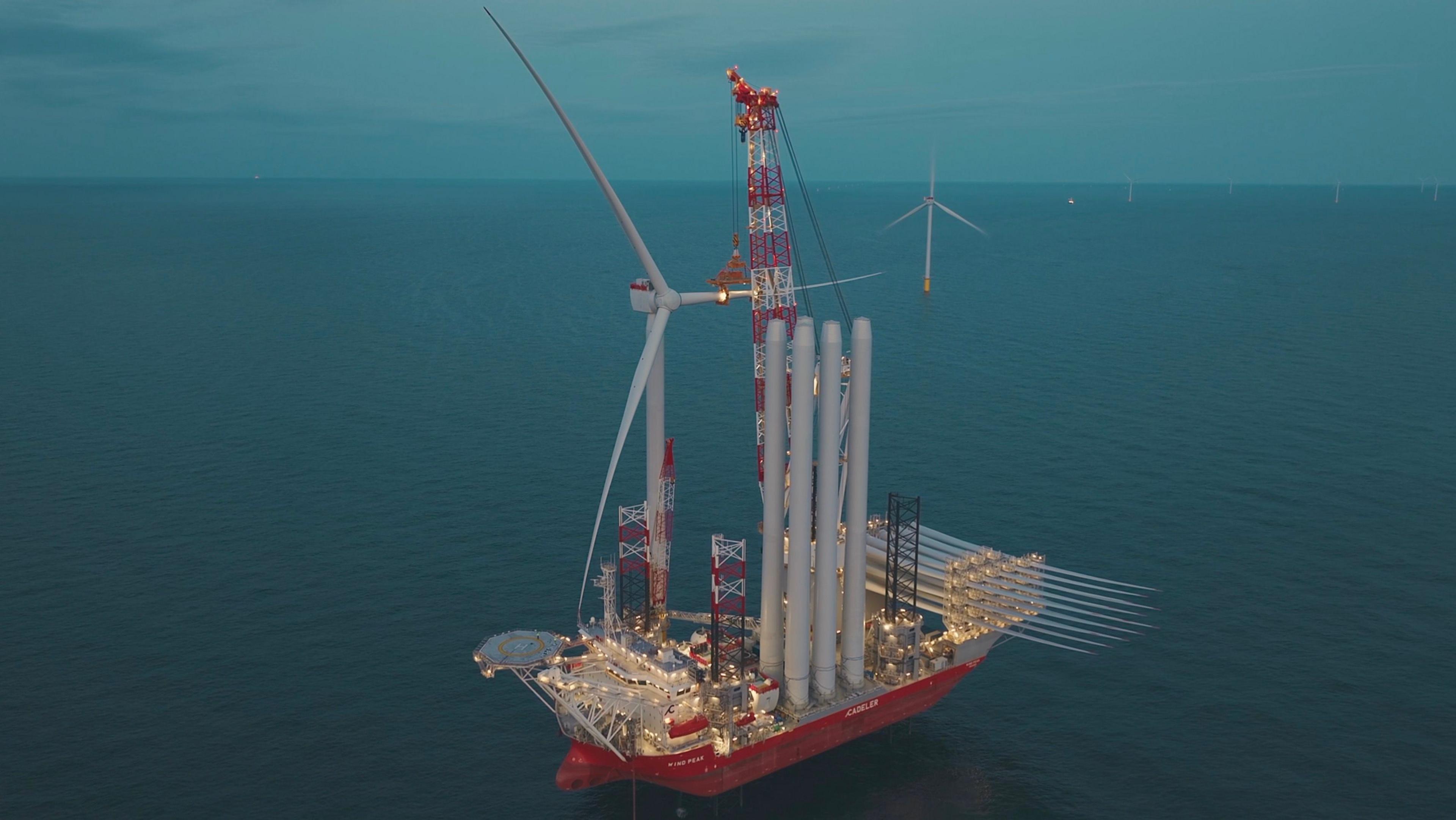A ship laden with wind turbine components is at sea. An assembled wind turbine can be seen in the background.