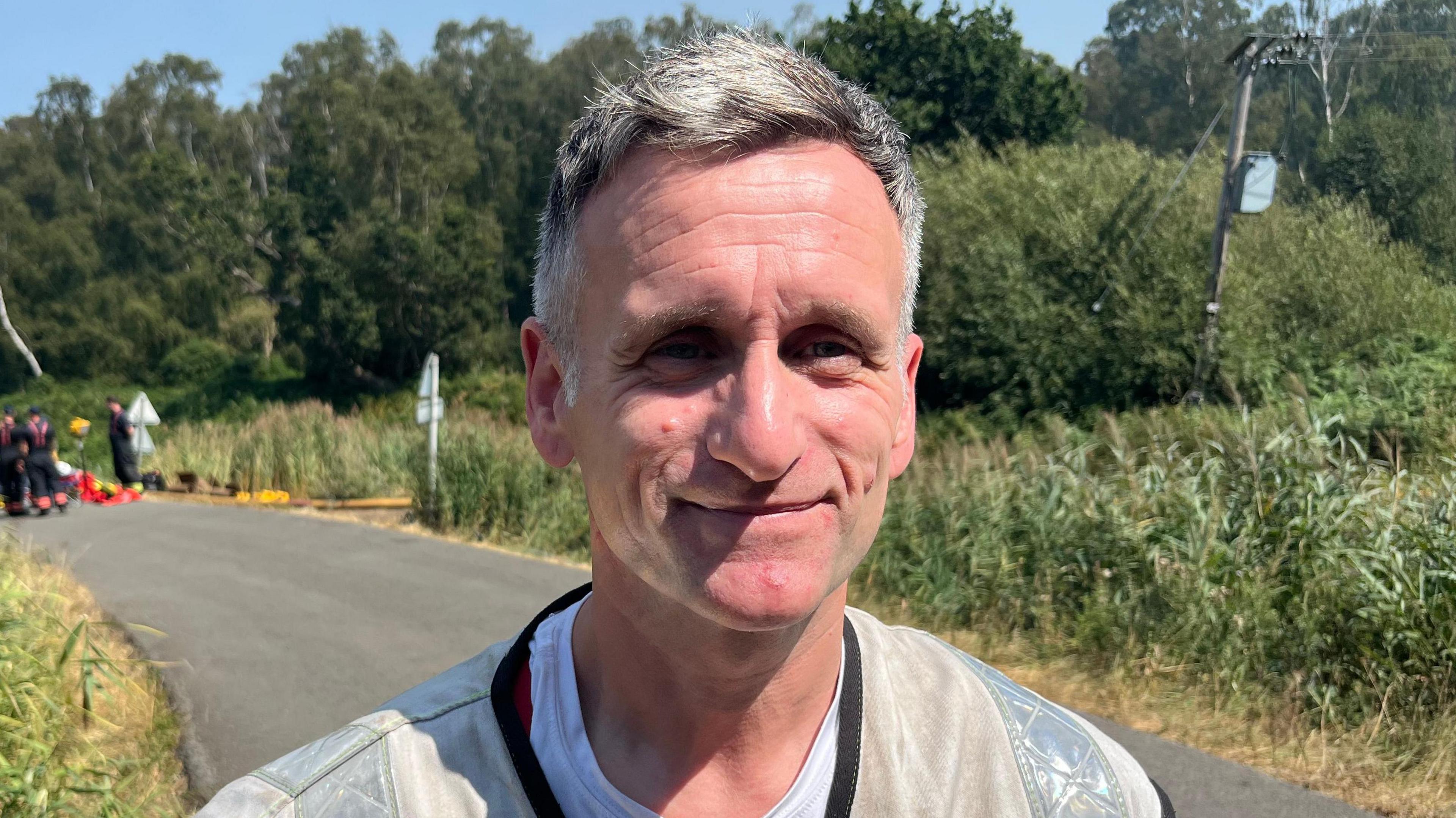 Wayne Swales has short grey hair. He is wearing a white shirt and a silver Hi-Vis jacket. He is standing on a road next to woodland.