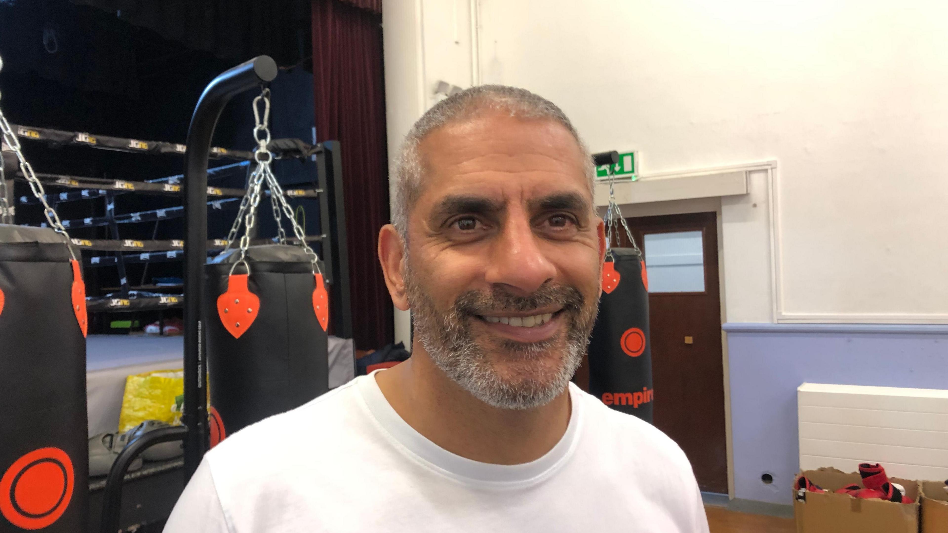 Paul Gill is wearing a white t-shirt. He has cropped grey hair and a beard and moustache. Behind him is a boxing ring on a stage. Punch bags hang on stands on the floor below.