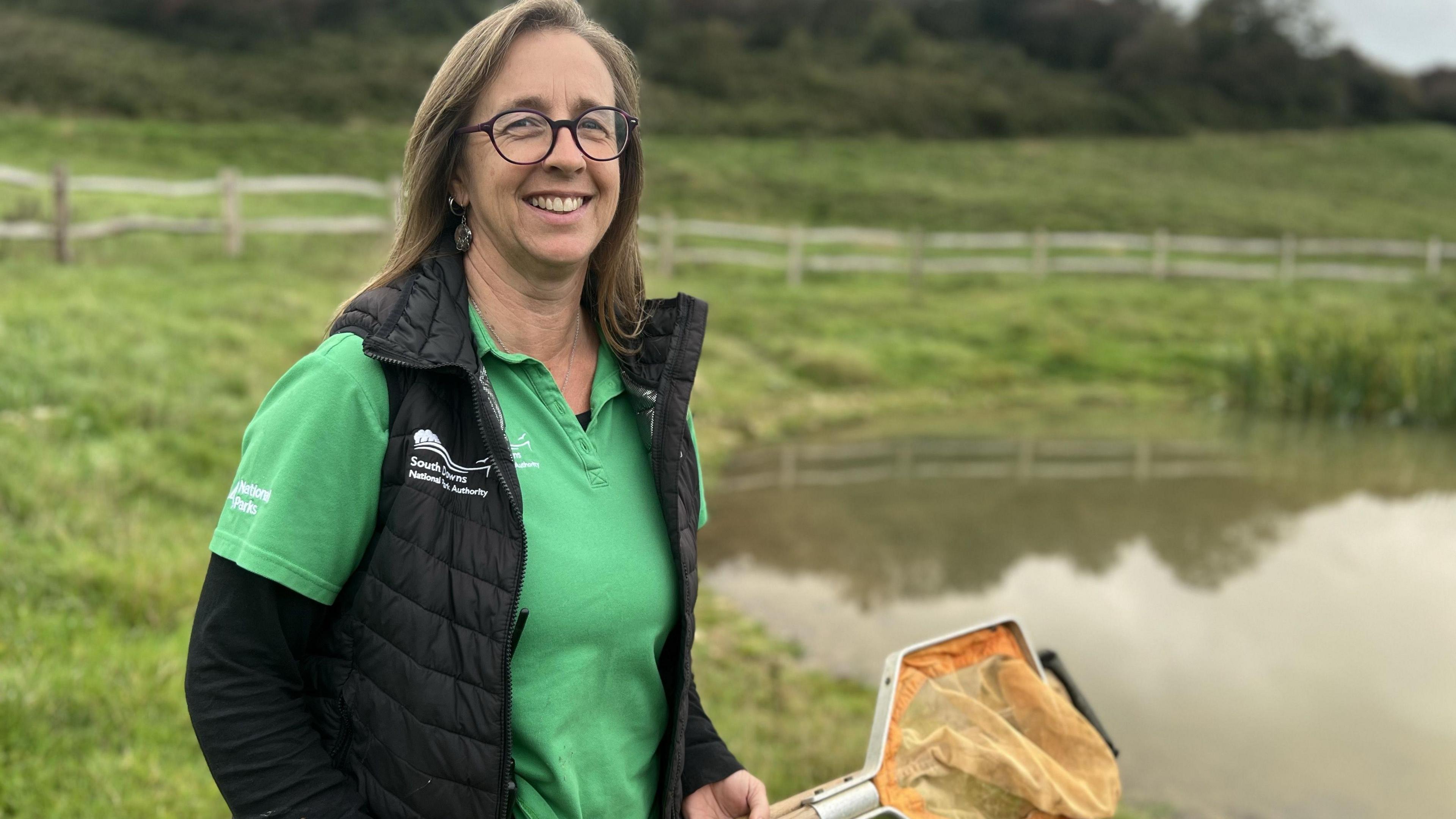 A woman with long brown hair and glasses smiles at the camera. She is wearing a green polo shirt and a black gilet jacket, She is holding an orange net. In the background you can see a brown pond.