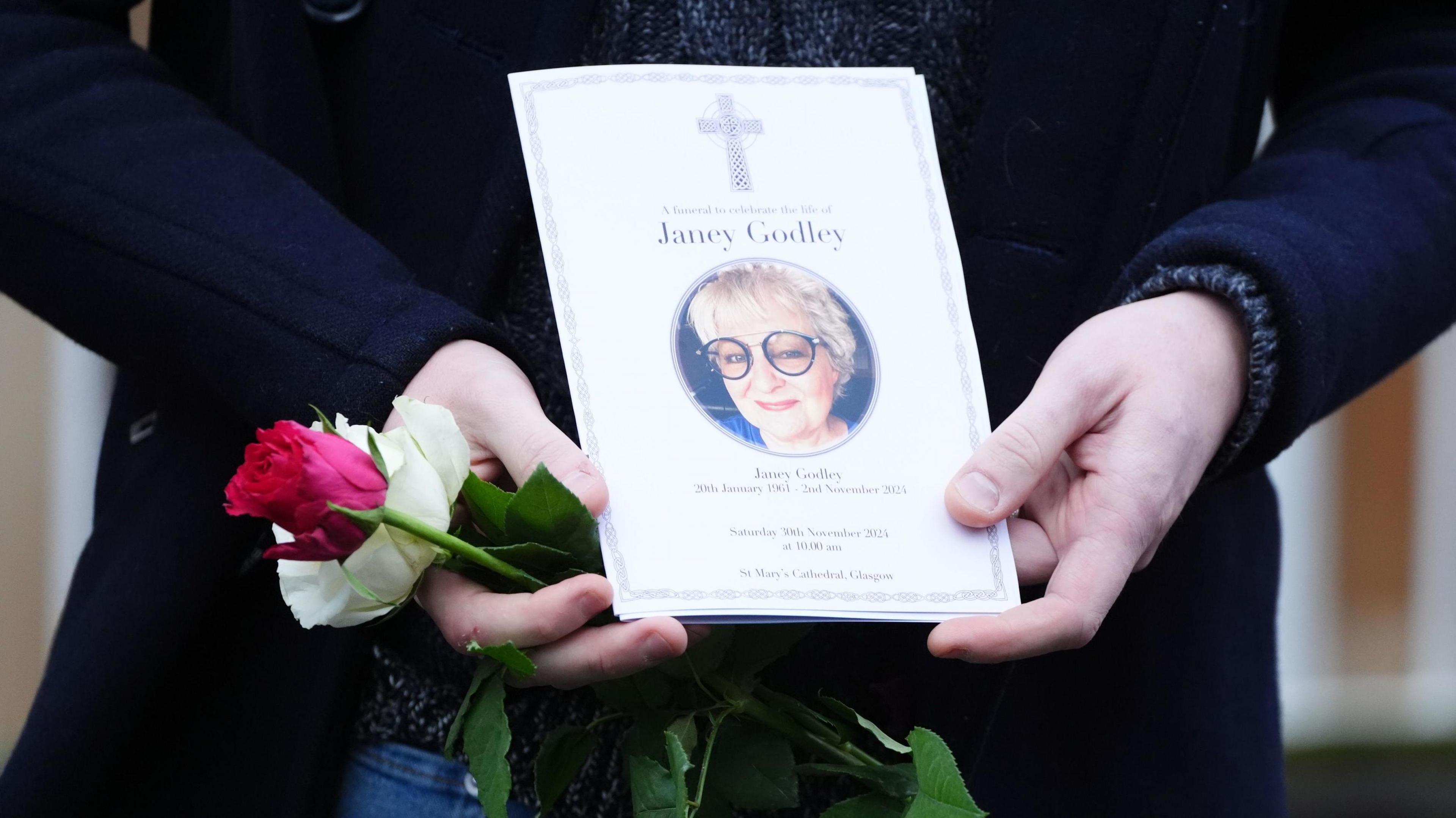 A mourner in dark clothing in close upholding a red rose and an order of service. We can only see the person's lower torso. The order of service names Janey Godley beside a photo of the comic wearing a large pair of round sunglasses.