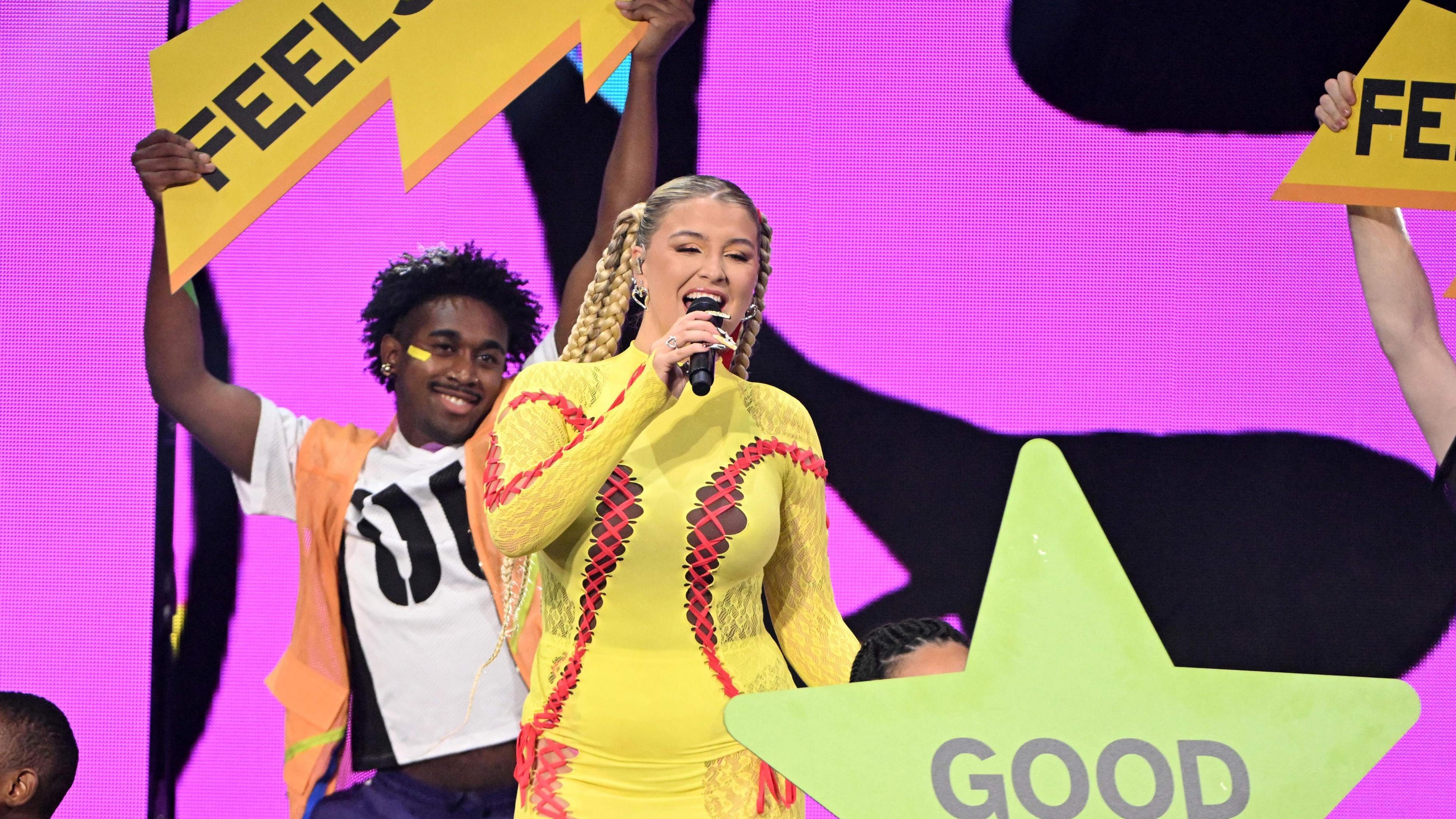 Caity Baser on stage. She wears a yellow two-piece with red laces across the sleeves and front of her top. She has her long blonde hair tied back in plaits and dancers around her hold up green and yellow signs reading 'feels' and 'good'. 