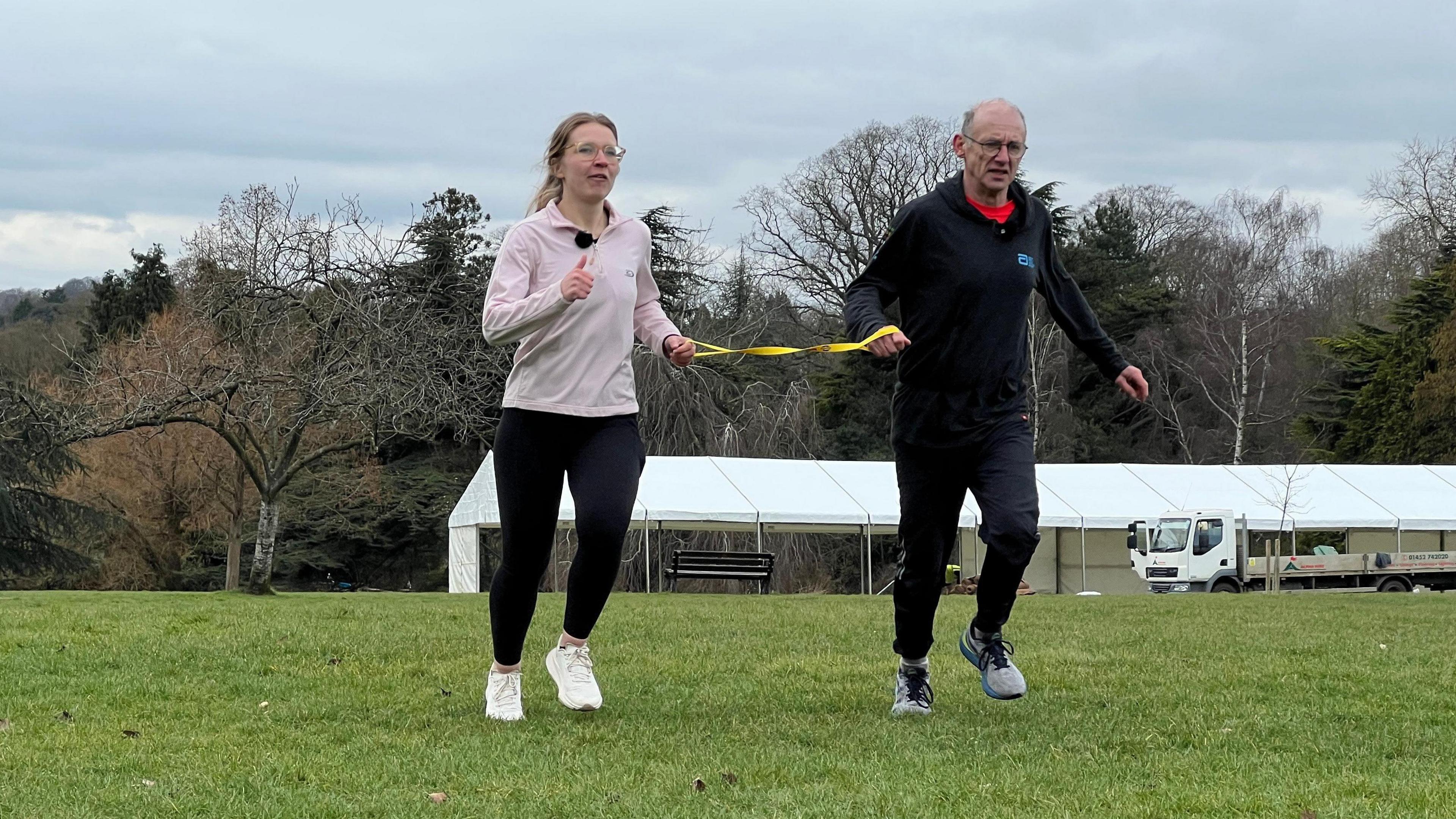 Karolina Pakenaite running with her guide Tom Hutchison. They are held together with a yellow strap and running on grass. Karolina is wearing leggings and a pink top and white trainers while Tom is dressed in a black tracksuit. They are running on grass. 