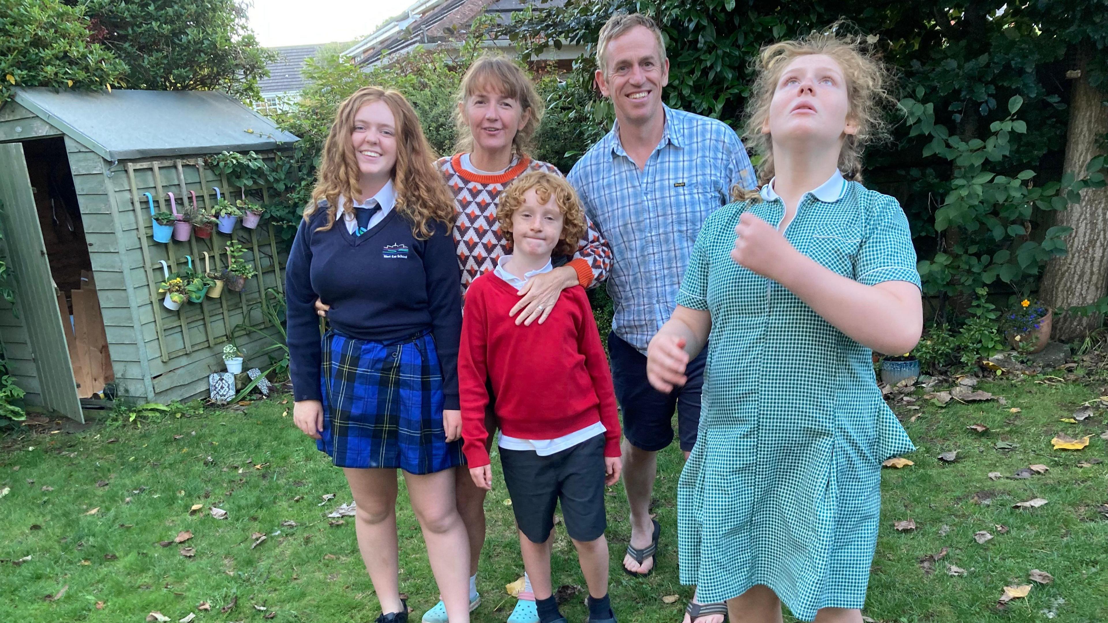 A family in their garden. On the right is a teenage girl in green and white checked school dress - she is looking upwards. Next to her are her mum and dad and her younger brother and older sister.