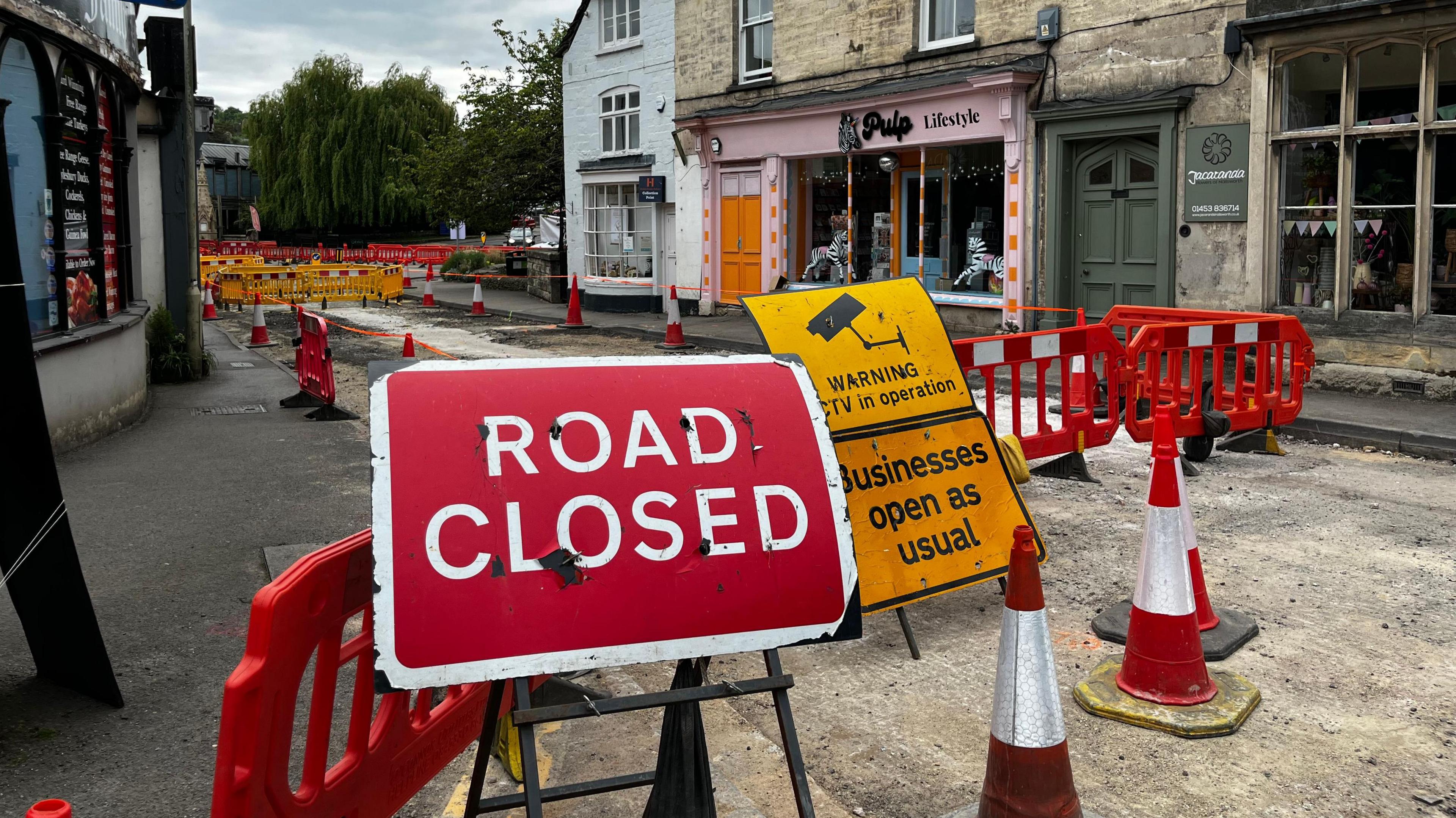 A large red Road Closed sign is next to a number of traffic cones and red bollards indicating that the road is closed. The road surface has been taken up which is why the road is closed and the shops look empty.
