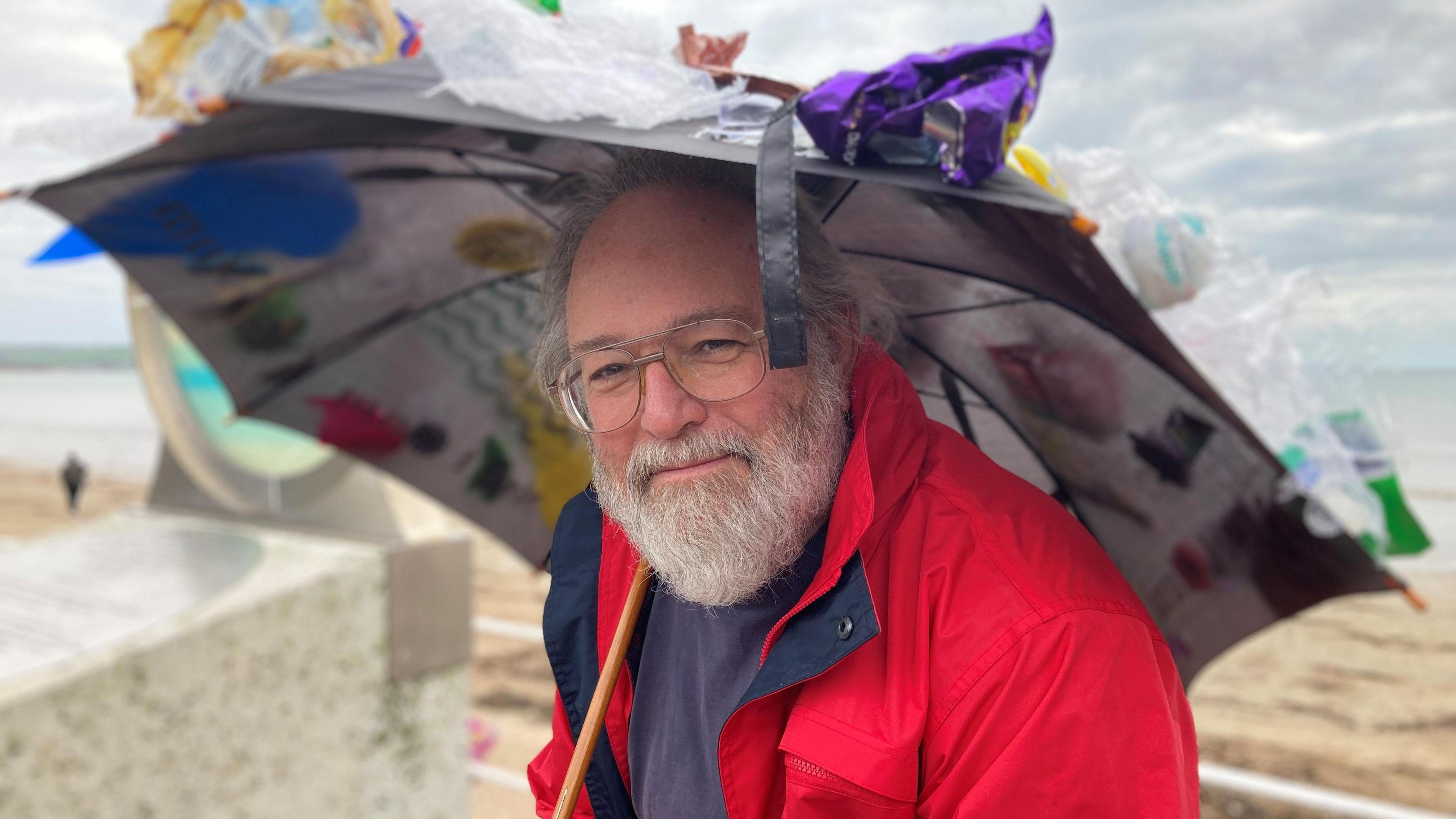 A man with white hair and a white beard, wearing a red coat, a blue top and a pair of glasses, holding an umbrella and smiling at the camera.