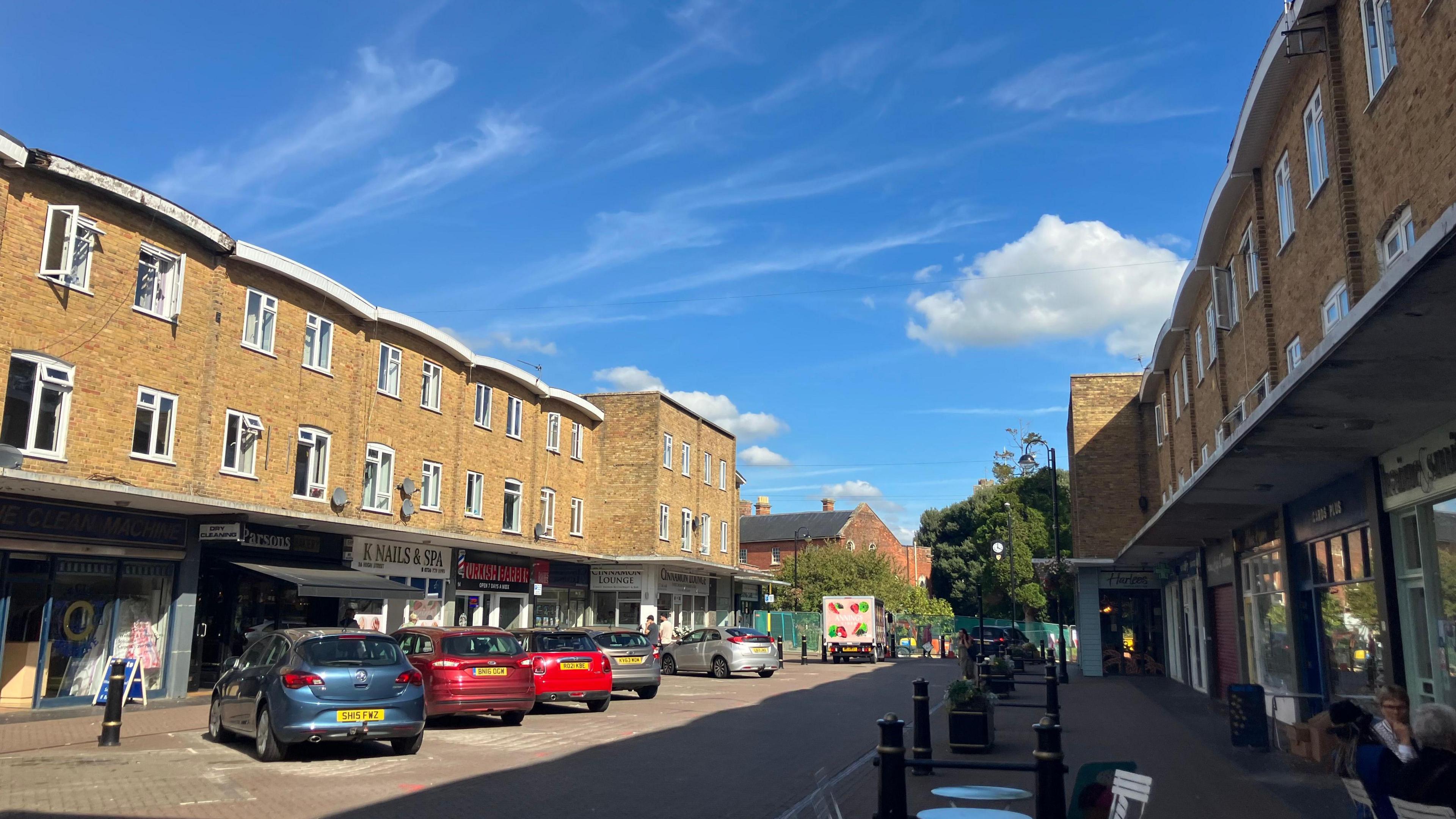 A view down Westbury high street - 1960s-style three storey buildings on either side, shops in the bottom and some cars parked down the left hand side. A sunny day.