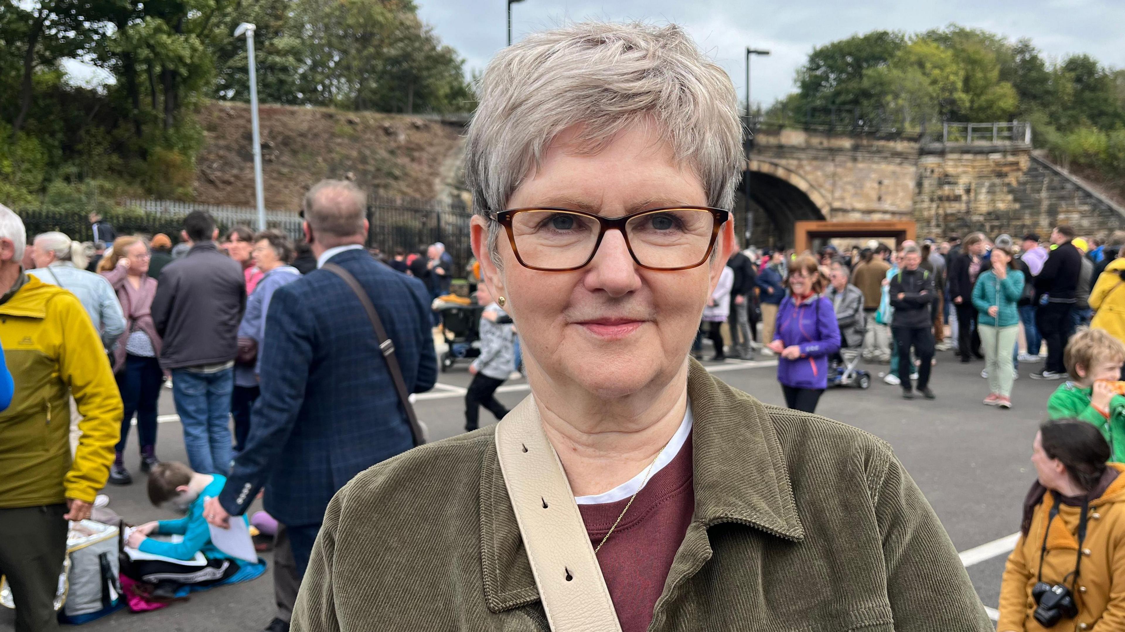 Head and shoulder view of an elderly woman, with short grey hair and spectacles. She is wearing a light green corduroy jacket over a maroon jumper. Behind her people can be seen milling around in front of a railway embankment and stone bridge.