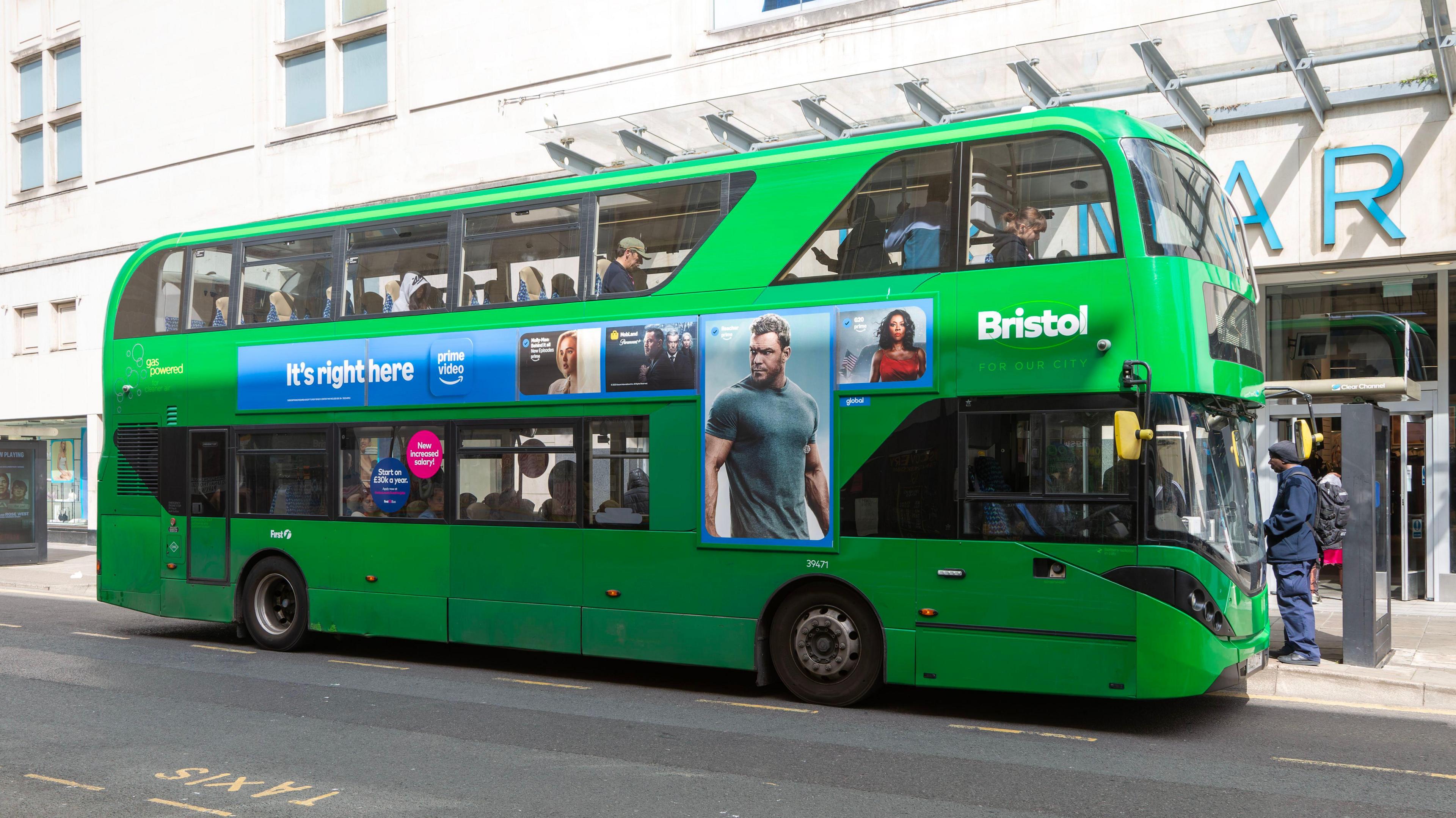 A green double decker bus at a bus stop outside Primark in Bristol city centre. There is a person standing at the stop waiting to board.