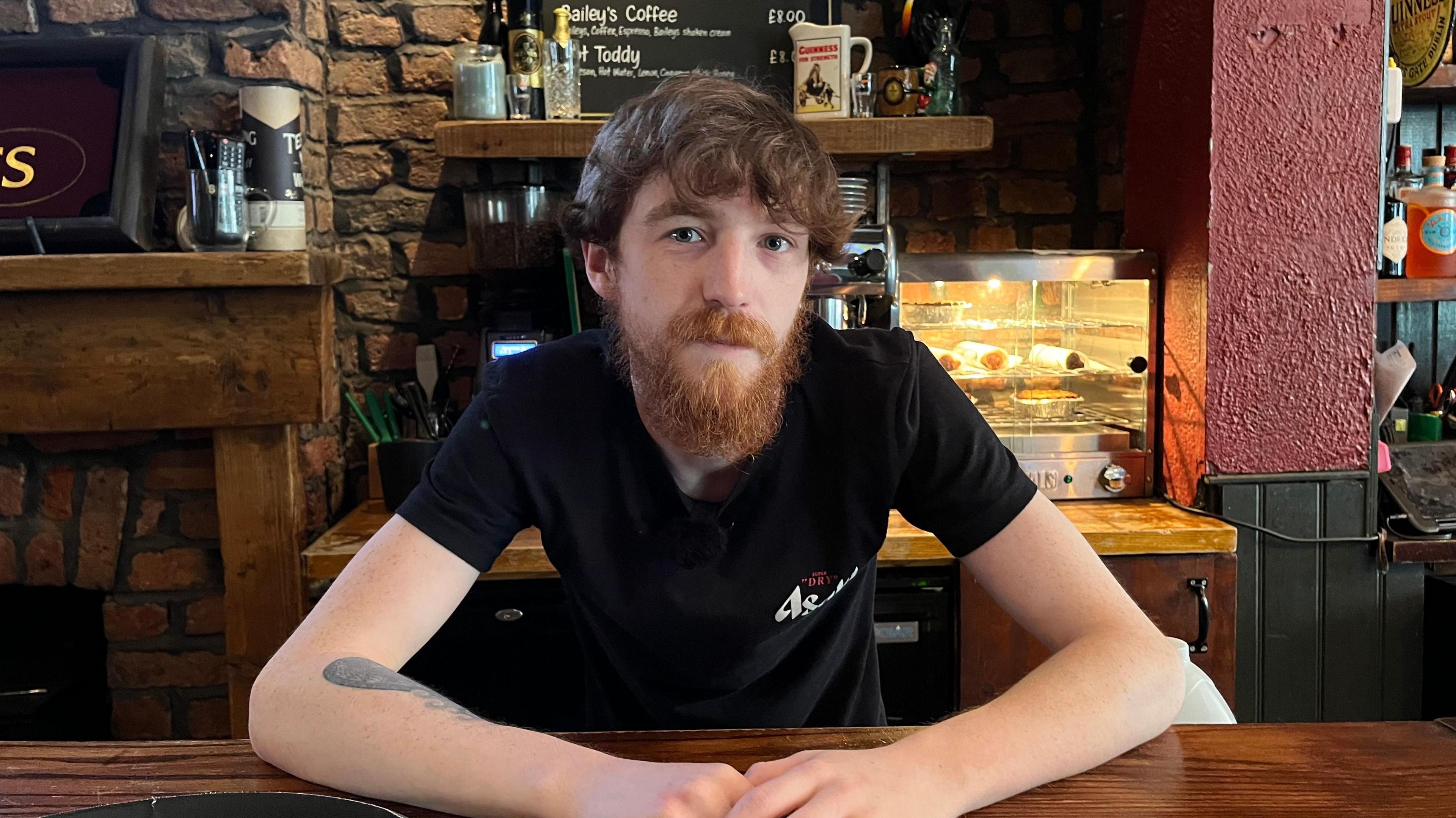 A man sits at a wooden bar inside a traditional pub, with a drinks menu, brick walls and shelves of bottles in the background.