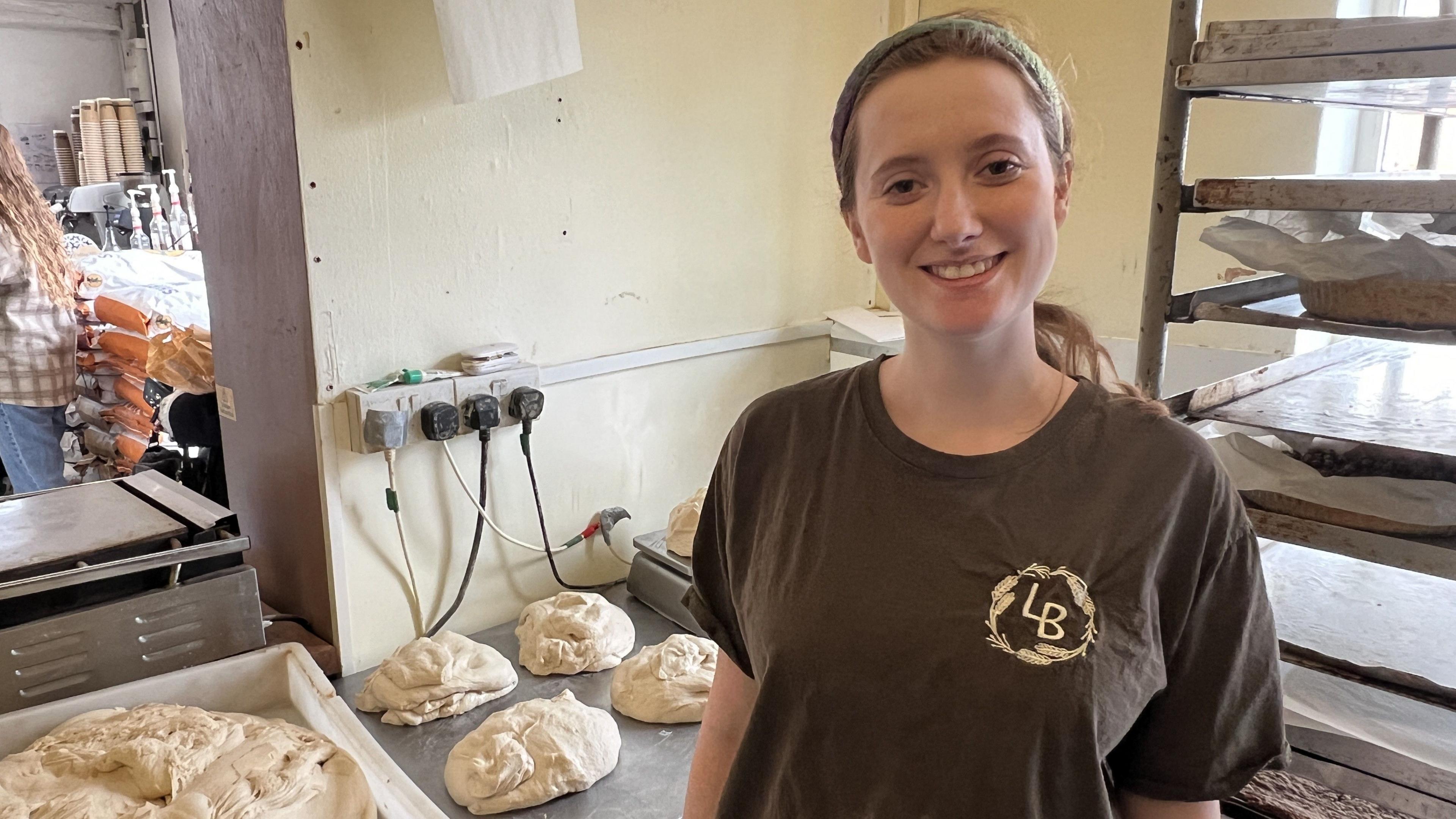 Sarah Rodborough wearing a dark brown t-shirt with a circular wheat logo which says LB in the middle. She has long brown hair which is pulled back and is wearing a green headband. She is standing in the Little Bakery, smiling at the camera. Behind her are metal sheets with piles of raw dough on them. On her right are metal shelves with cooked loaves of bread stacked up on baking paper. 