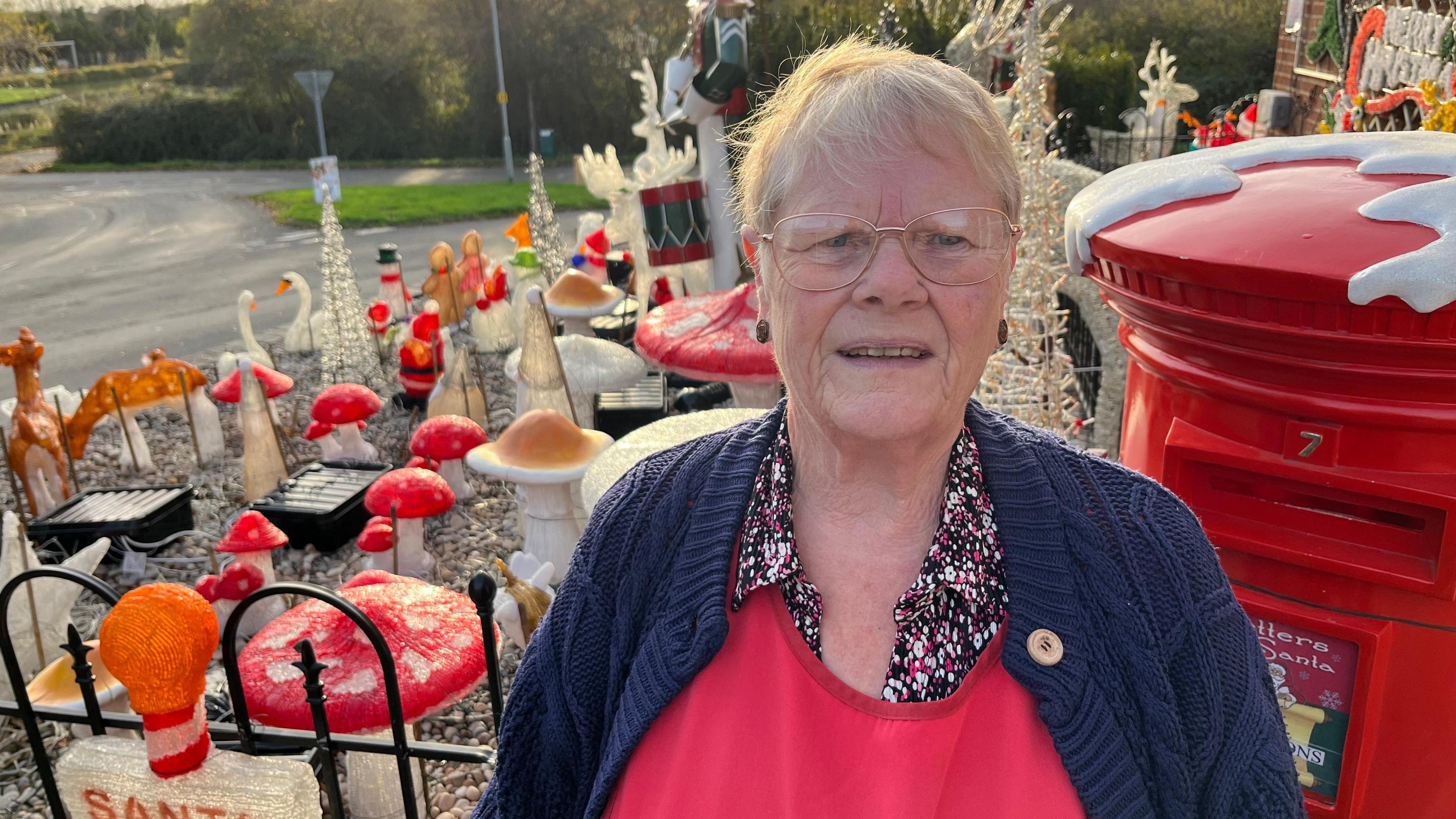 Helen Attlesey is looking at the camera and is standing in front of lots of Christmas decorations in a garden. There is a postbox, and decorations including swans, deer, toadstools and snowmen. She is wearing a patterned blouse, a red top and a blue cardigan. She has short, fair hair and is wearing spectacles.