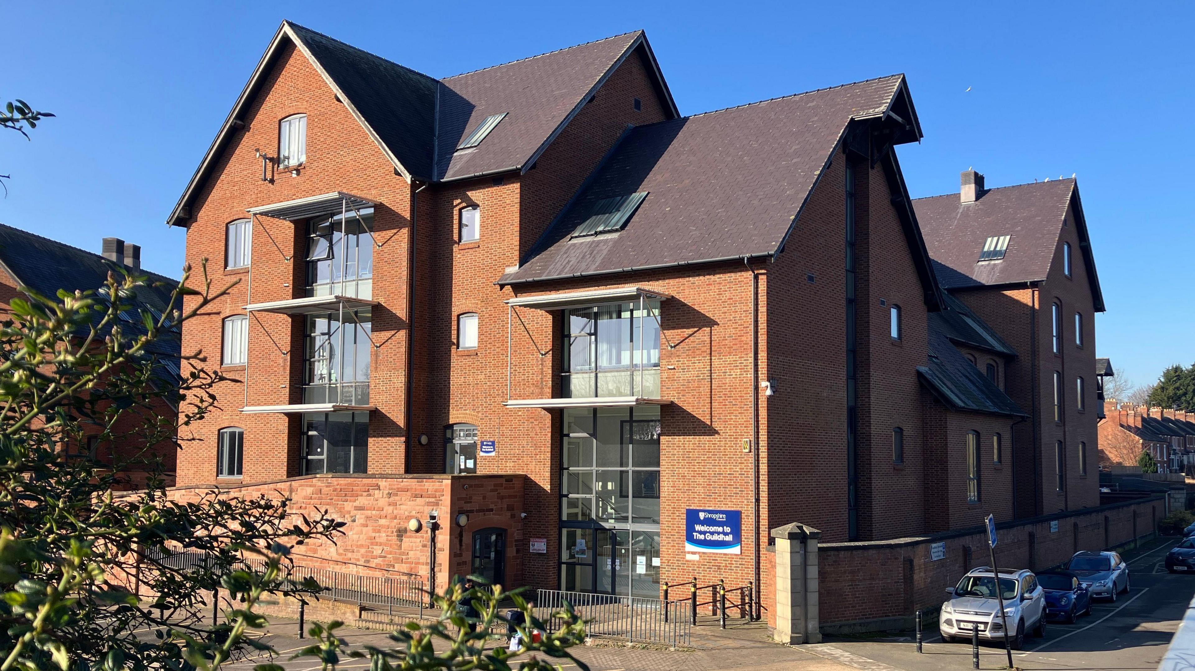 A five-storey red-bricked council office with various pitched roofs and gable-end walls. The sky is blue and three cars are parked in the shadowed side of the building.