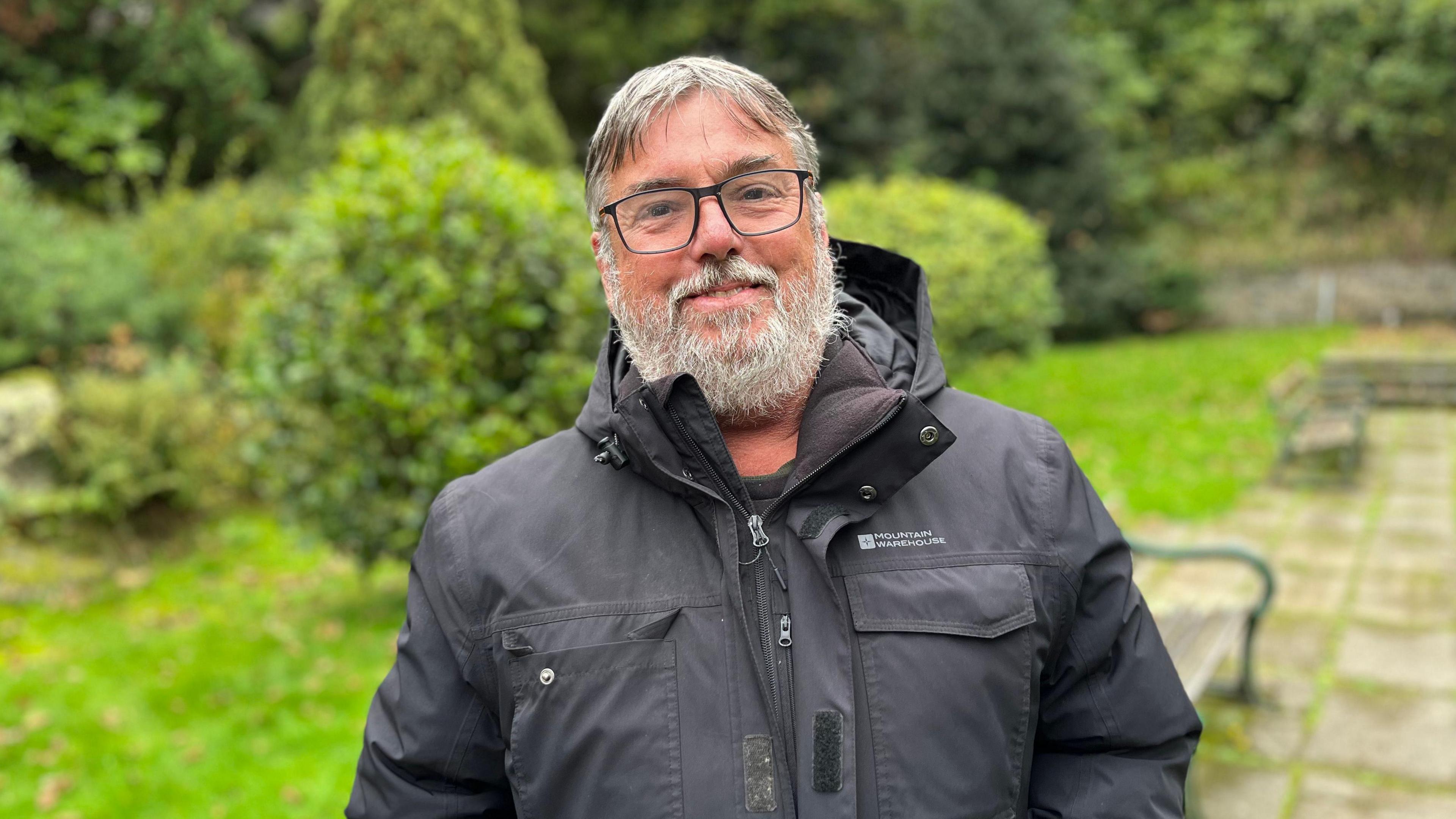 A man with grey hair and a grey beard looking towards the camera wearing black glasses. He's wearing a black coat.