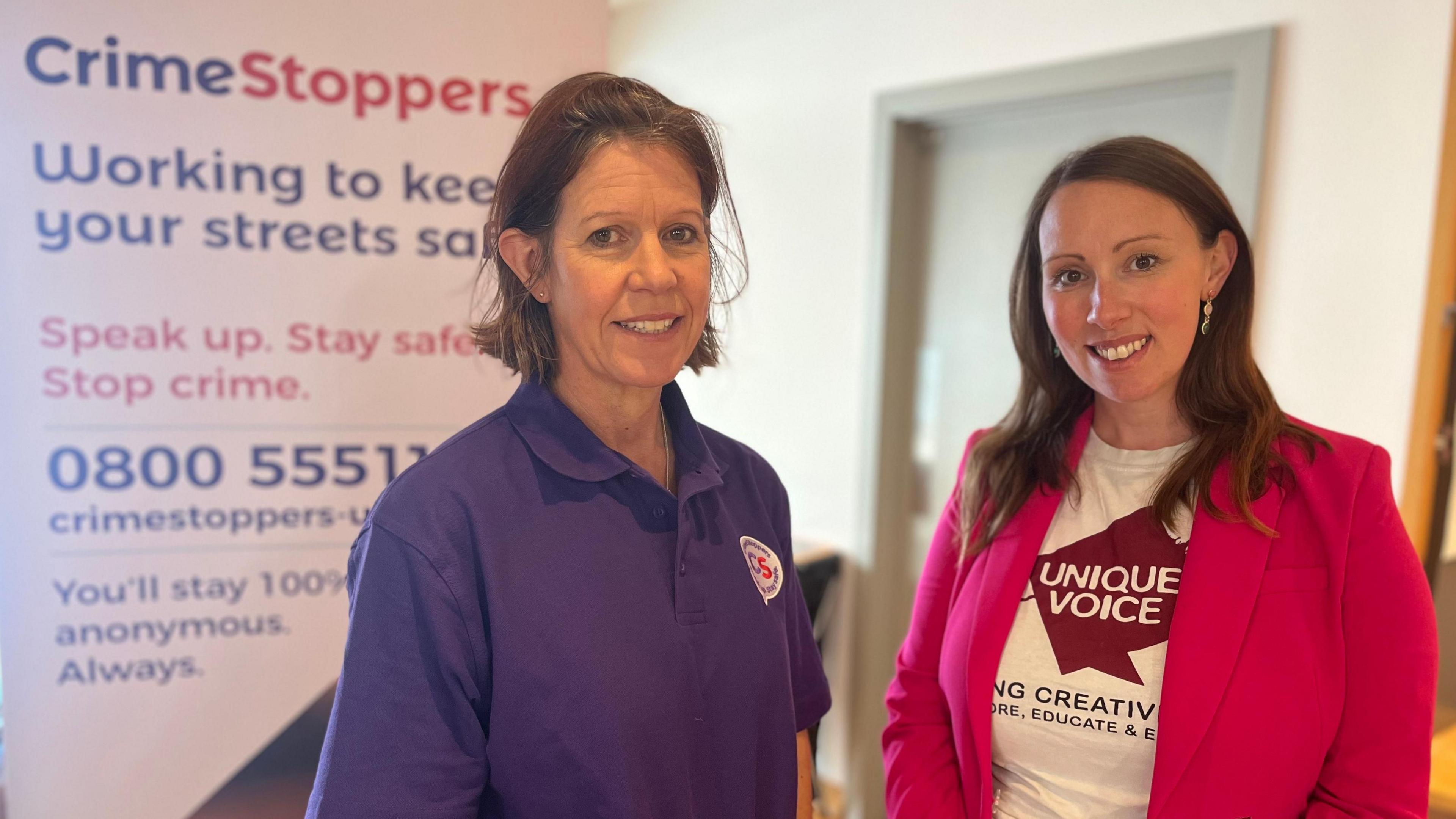 Karen MacDonald (left) from Crimestoppers in purple polo shirt and Krystal Keely from Unique Voice in pink blazer standing in front of a Crimestoppers banner inside a room