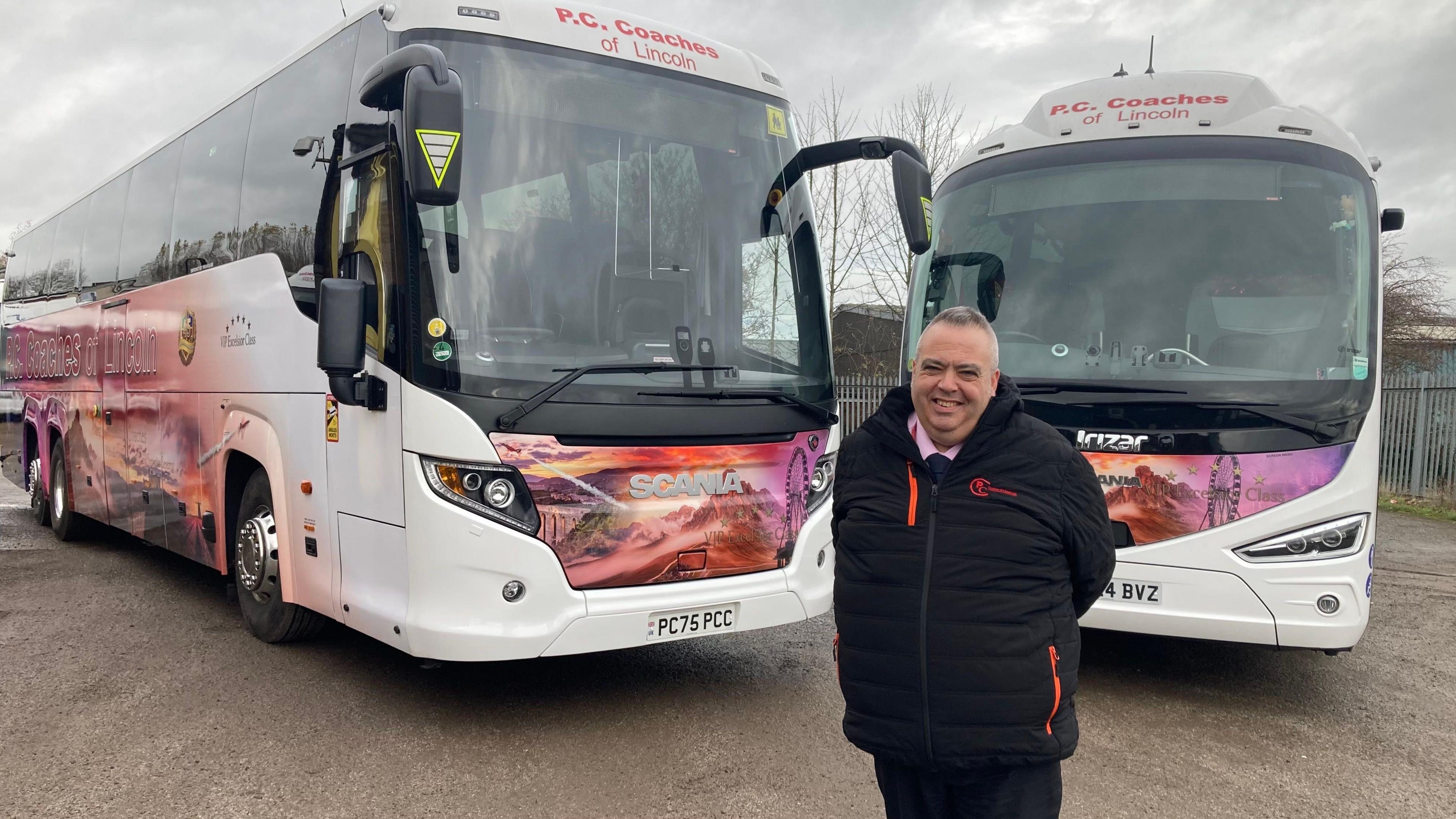 A man with short grey hair, dressed in a thick black coat, smiles as he stands in front of two large coaches with big tinted windows. The bodywork is partially white and partially painted in shades of red and purple with scenes of holiday locations including mountains and ferris wheels.