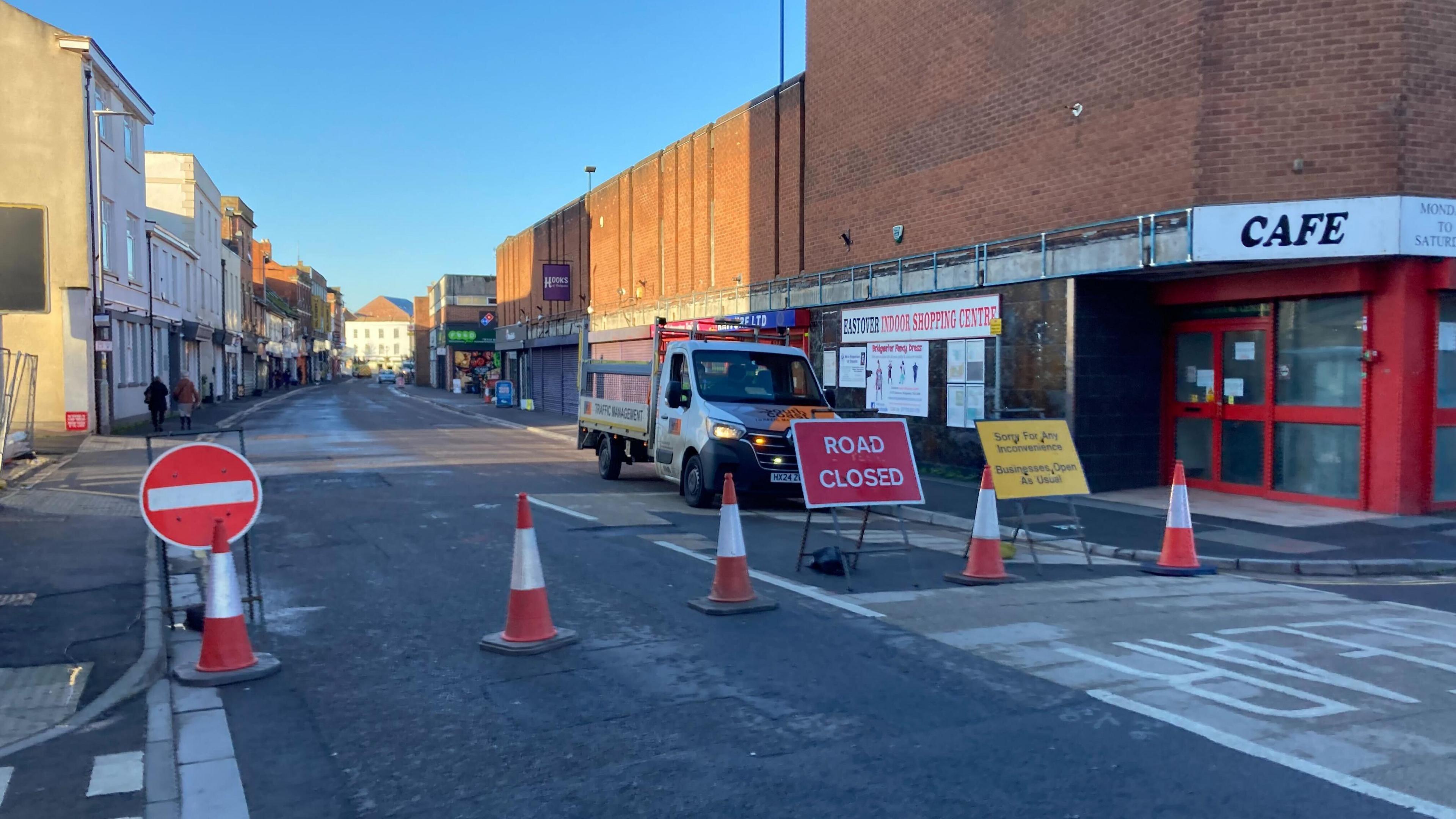 Traffic cones block a shopping street, with a work van parked behind them. A sign reads "road closed". There are some people visible on the pavement next to the shops, and it is a bright sunny day. 