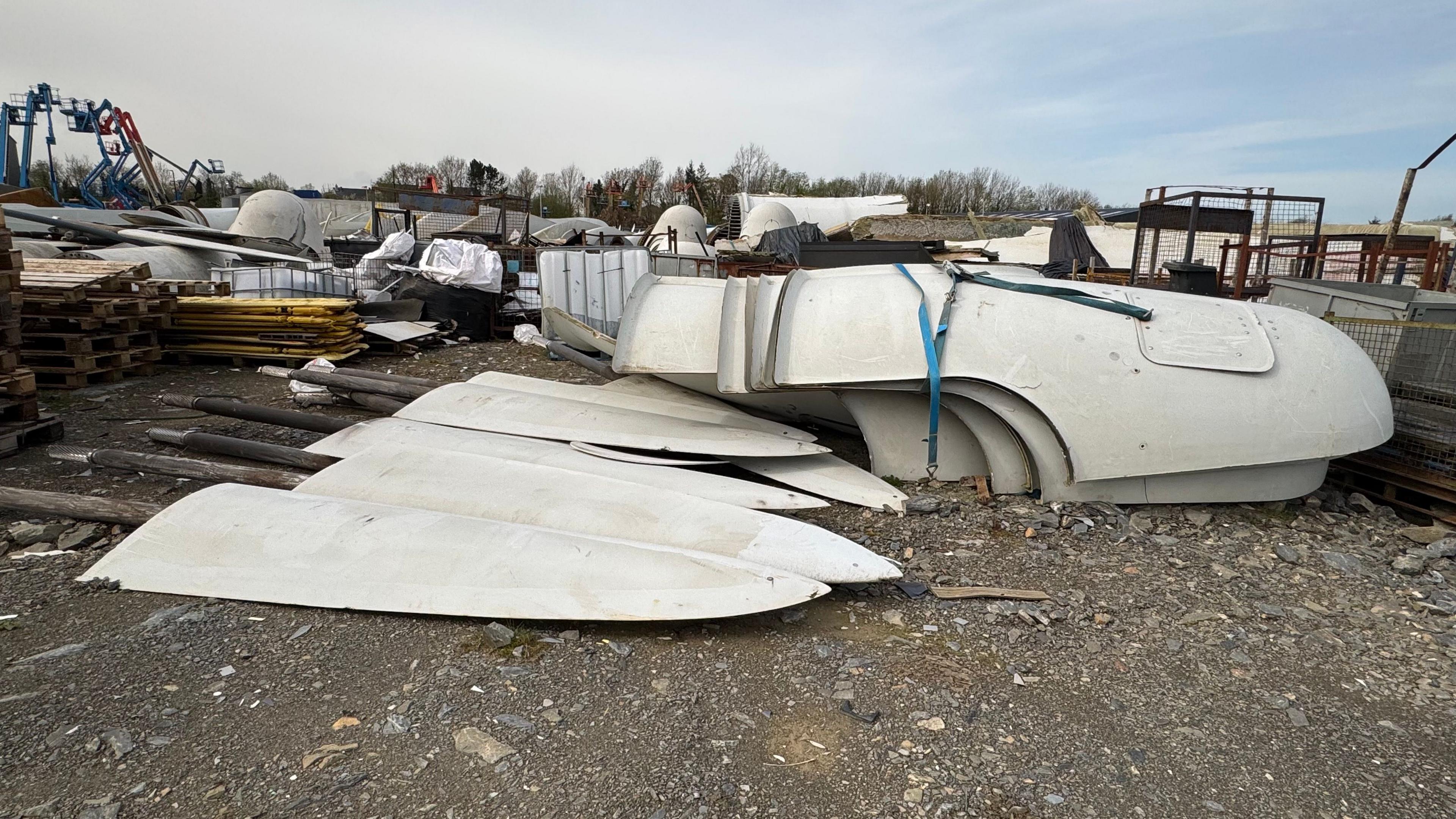 Large piles of disused turbine waste is piled up in an industrial yard.  It includes at least six large white tips of turbine blades.  