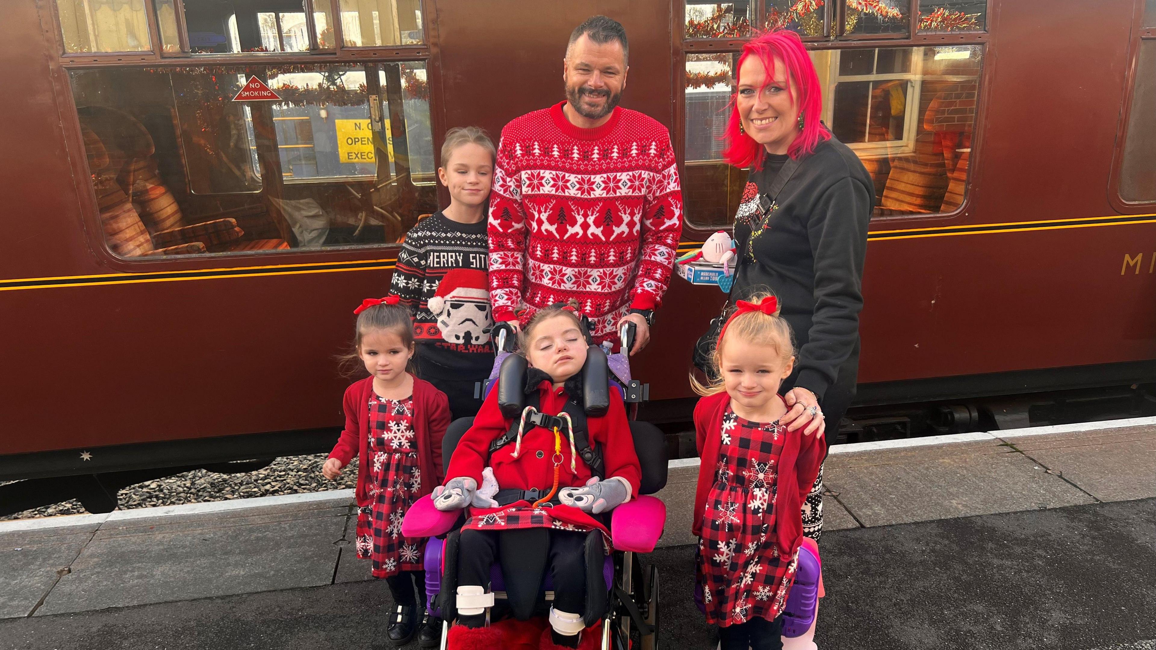 Three children are at the front and at the back is a boy on the left, a man in the middle and a woman on the right. The man with dark hair and a beard is wearing a red and white jumper. The woman has red hair and is wearing a dark top.
