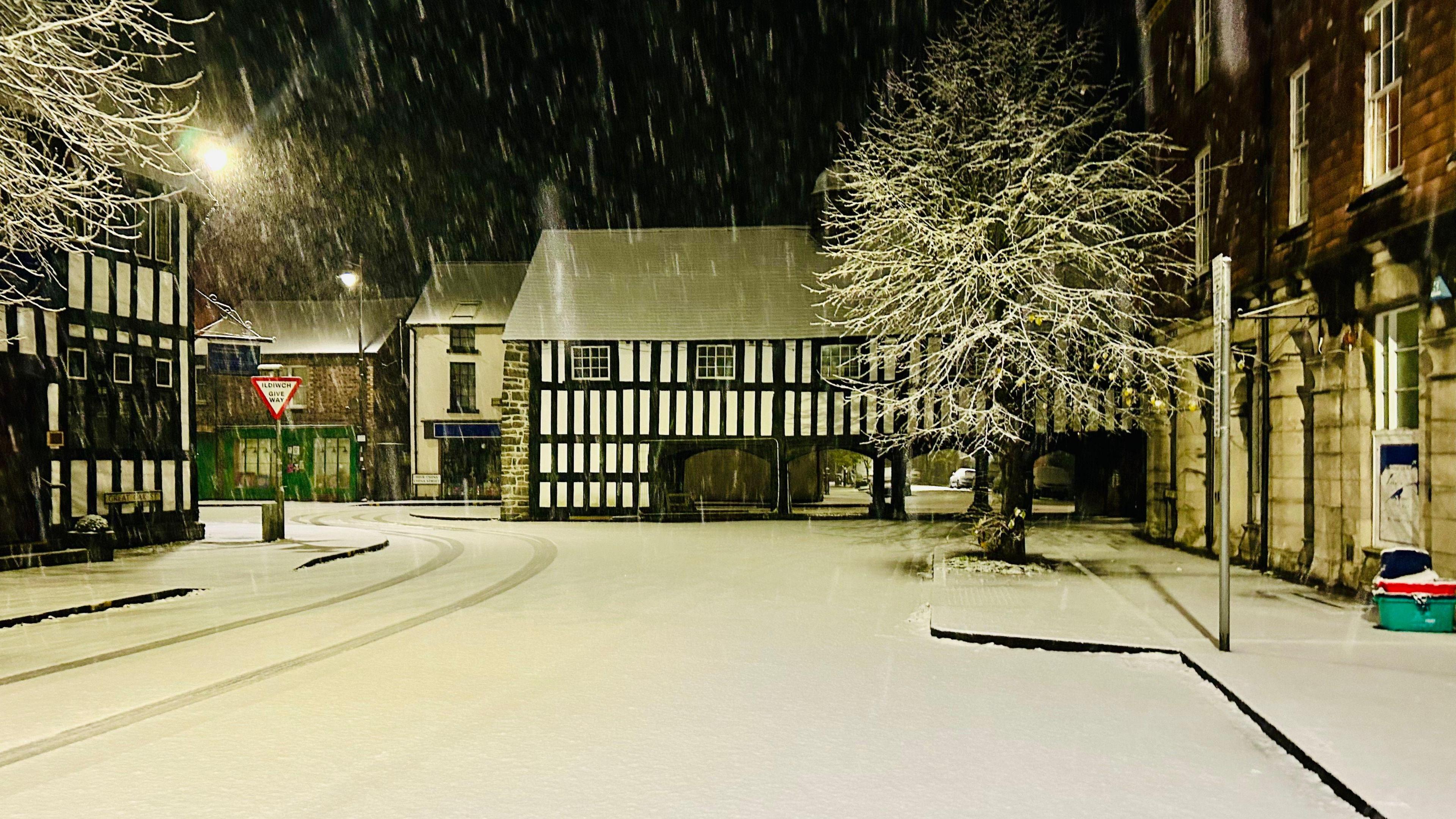 Road covered in snow in the town with Tudor buildings lining the road. The houses' rooves are covered in snow and more snow is falling