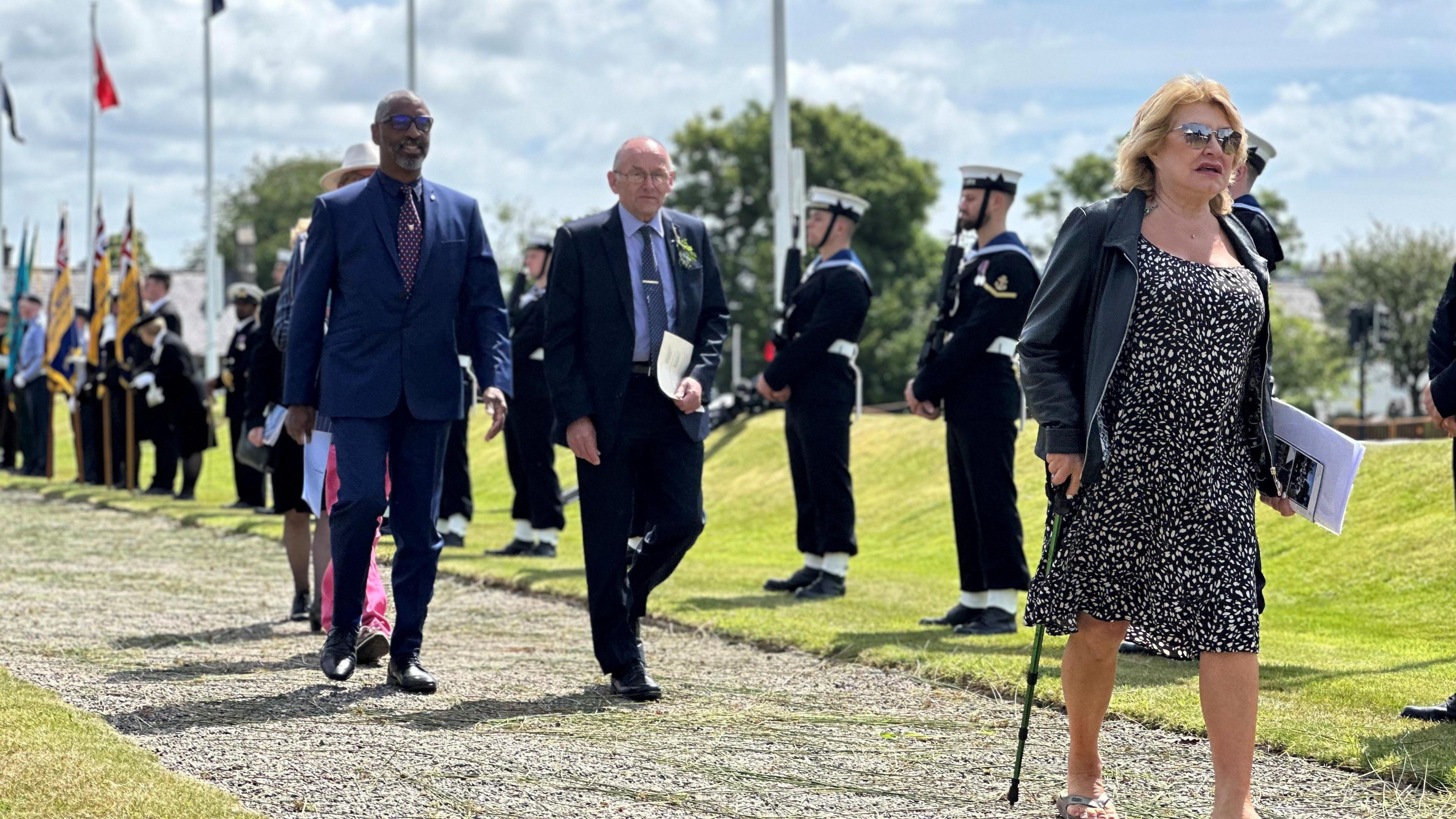 Smartly dressed petitioners walk along the ceremonial walkway. A woman wearing a black and white dress and black jacket and using a walking stick is in front of two men dressed in blue suits and wearing ties. The guard of honour and standard bearers line the walkway behind them. The walkway itself is covered in gravel and has green grass at either side.