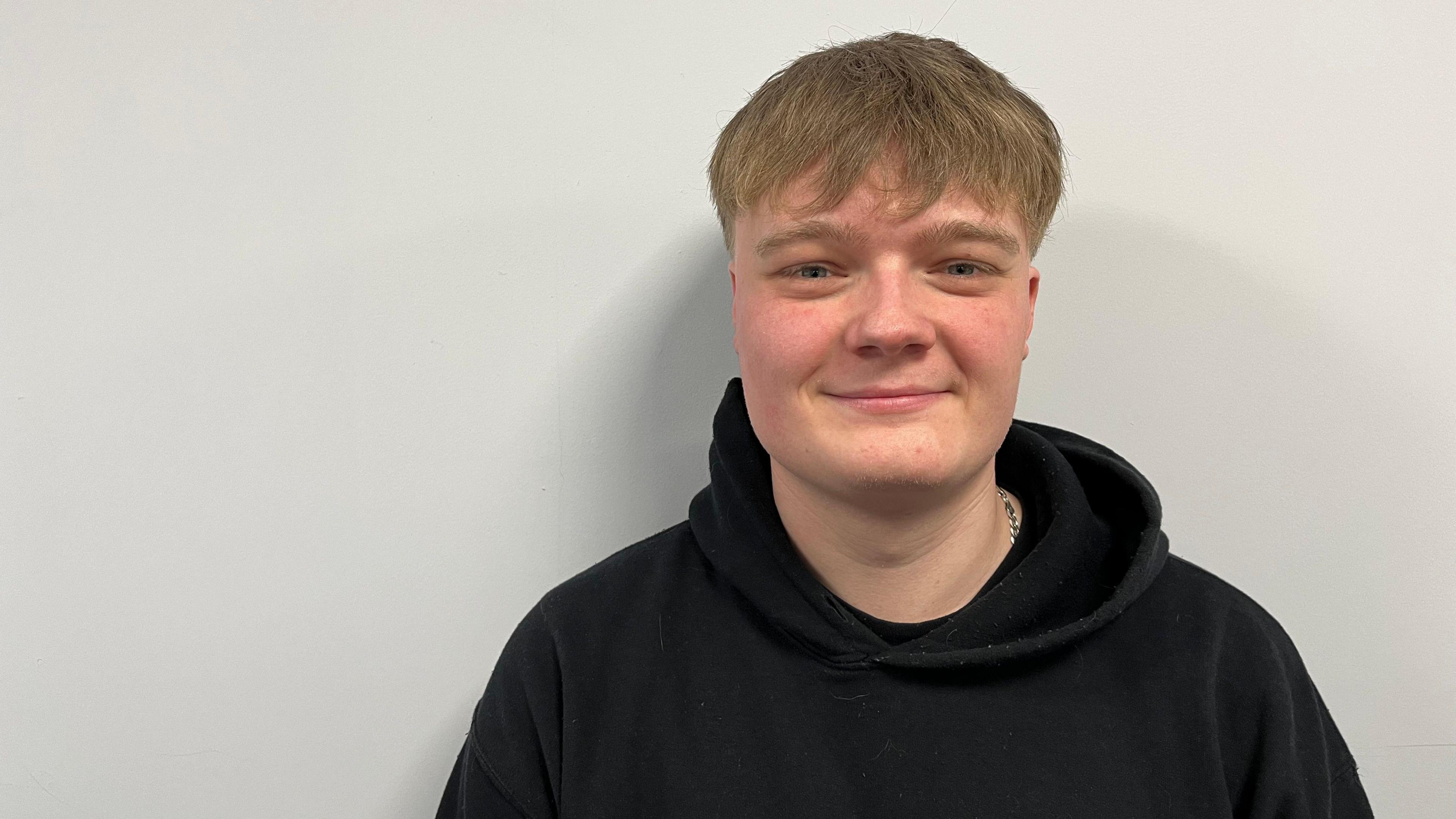 Jacob Shane with sandy coloured hair, wearing a black hoodie stands against a grey wall and smiles at the camera.